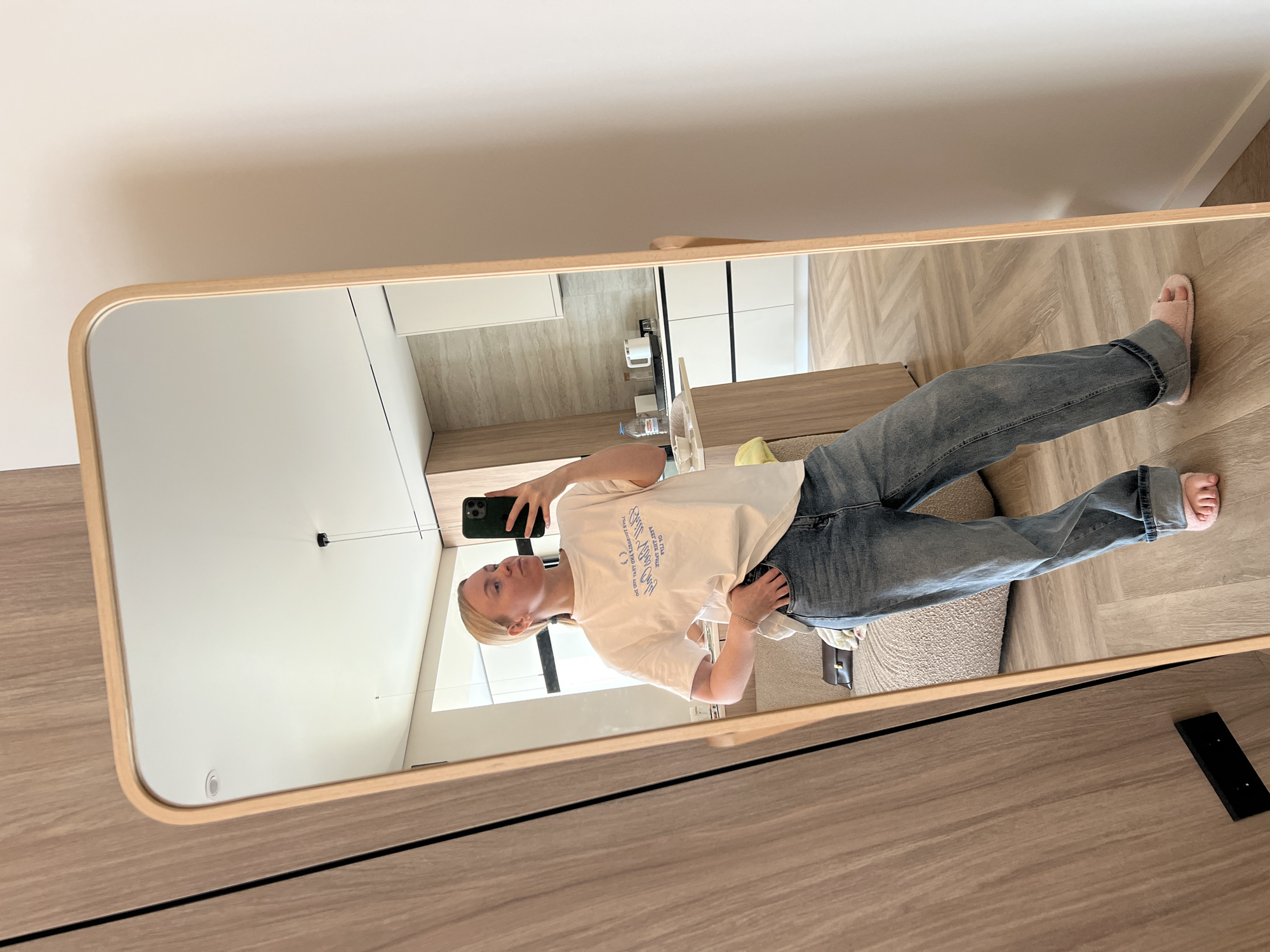 A young woman with blonde hair, wearing a white t-shirt with blue text and blue jeans, is captured in a mirror selfie in what appears to be a modern residential setting in Новое, Russia. She is holding a dark green smartphone, likely an iPhone, to take the picture, visible in the mirror's reflection. Her expression is neutral to slightly posed.
The setting reflects a contemporary interior, featuring light-colored walls and a light wooden floor laid in a herringbone pattern. In the background of the reflection, a kitchen area is visible with white cabinets and a light countertop or backsplash. A clear water bottle can be seen on one of the surfaces. Part of a dark piece of furniture, possibly a sofa or chair, is also reflected. The woman is wearing light pink fuzzy slippers.
Notable text on her t-shirt, written in blue, includes the phrases: "HAPPINESS IS FREE", "TO GET A HAPPY LIFE", "YOU MUST LOVE YOUR LIFE", "LOBSTER AND THE SEA", and "ON THE GENTLE WAVES". The time of day appears to be daytime, judging by the ambient lighting, though no specific light sources are prominent. The image offers no cues regarding the outdoor weather. The mirror itself has a light wooden frame, and the foreground outside the mirror shows more of the wooden floor and a dark, rectangular object, possibly part of the mirror's stand or another piece of furniture.