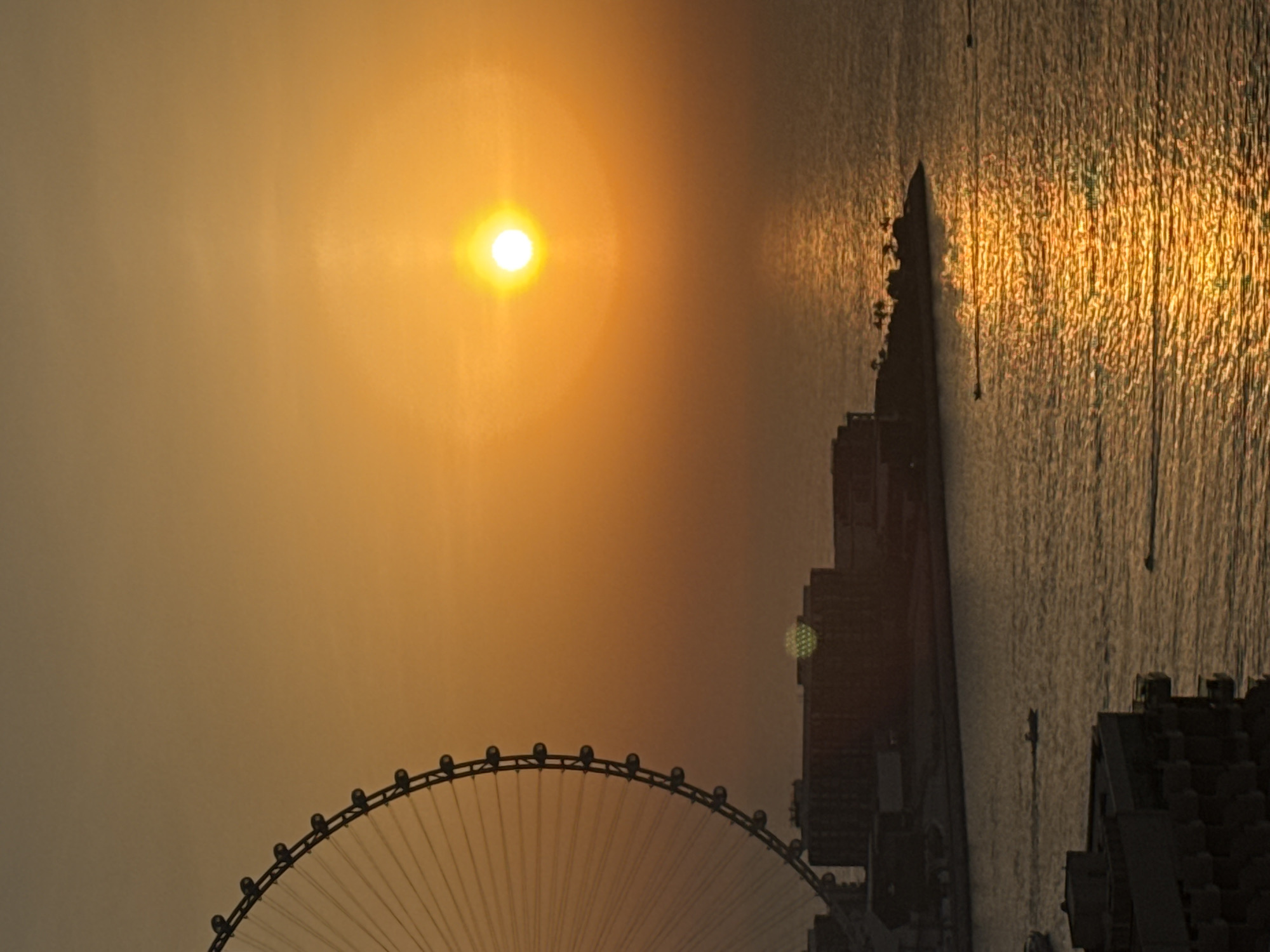 The image captures a sunset over the Arabian Gulf in Dubai, United Arab Emirates. The sun, a bright orange orb, is positioned in the upper right quadrant of the frame, casting a warm, golden glow across the sky and the water. The surface of the sea reflects the sun's light, creating a shimmering, rippling effect.
In the foreground, on the left side of the image, a portion of the Ain Dubai observation wheel is visible. This massive Ferris wheel, a prominent landmark in the city, is silhouetted against the vibrant sky. Below the wheel, a low-rise building complex is visible, with darker, more complex structures behind it.
On the water, several small boats are navigating the calm sea, leaving faint wakes in their paths. The overall impression is one of tranquility and natural beauty, with the man-made structures of Dubai integrated into the serene coastal landscape. The lighting suggests either early morning or late evening, with the warm tones indicating a sunset or sunrise.