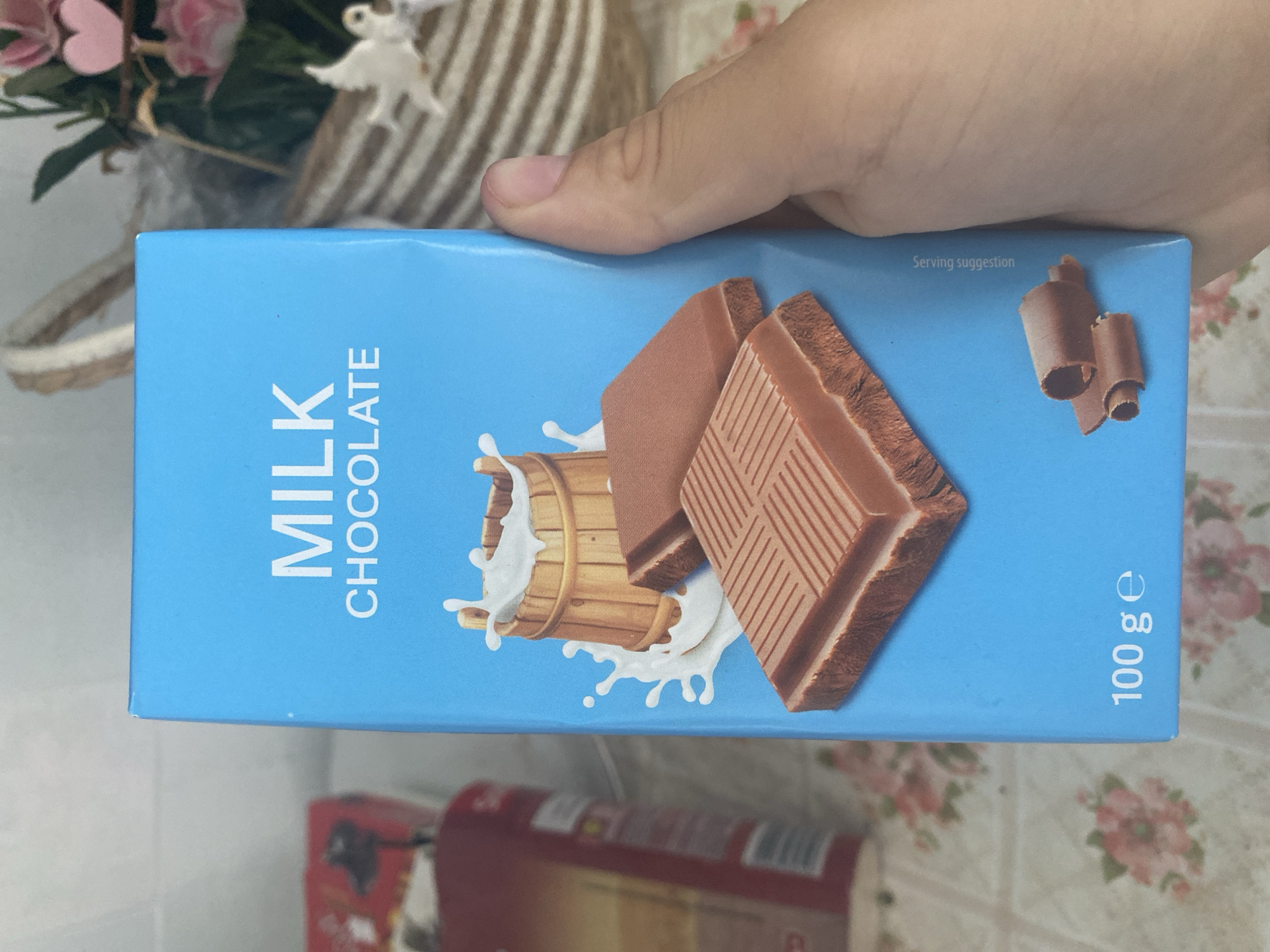 In Blagoevgrad, Bulgaria, a person is holding a blue cardboard box of milk chocolate. The box features an illustration of a wooden bucket with milk splashing out, and two pieces of milk chocolate. The text on the box reads "MILK CHOCOLATE" and "100 ge". The background shows a table with a floral tablecloth and a basket with flowers and decorative figures.