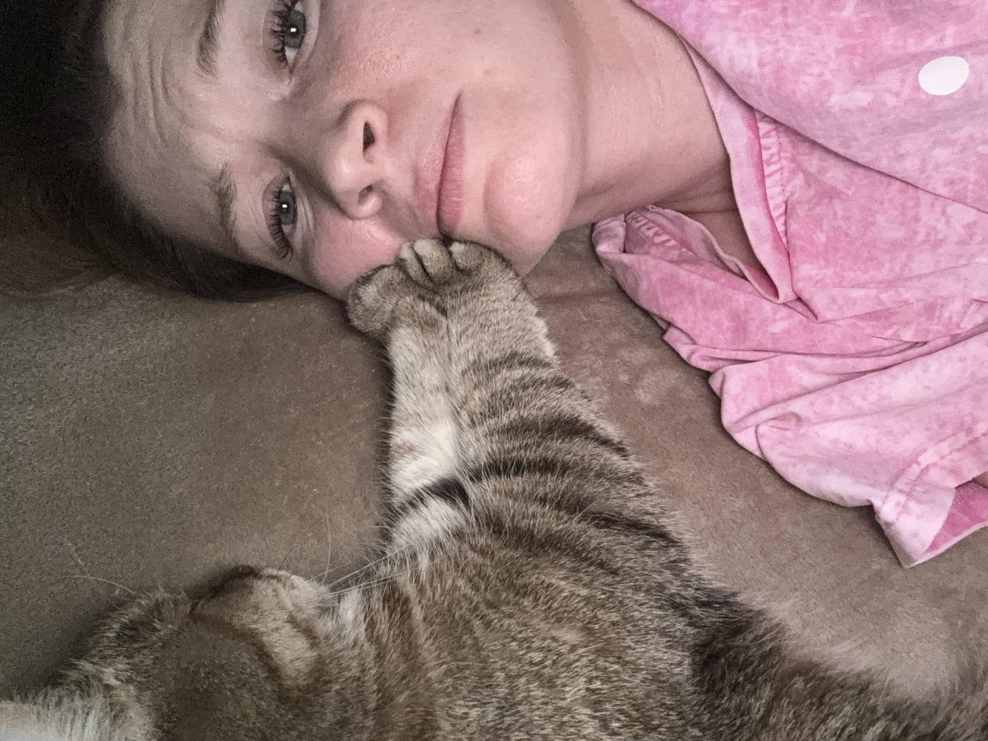 A woman lies on a brown surface, possibly a bed or couch, wearing a pink shirt. A tabby cat is positioned near her face, with its paw touching the woman's face. The scene appears to be indoors. No information suggests this image was taken in Chaikovsky, Russia.