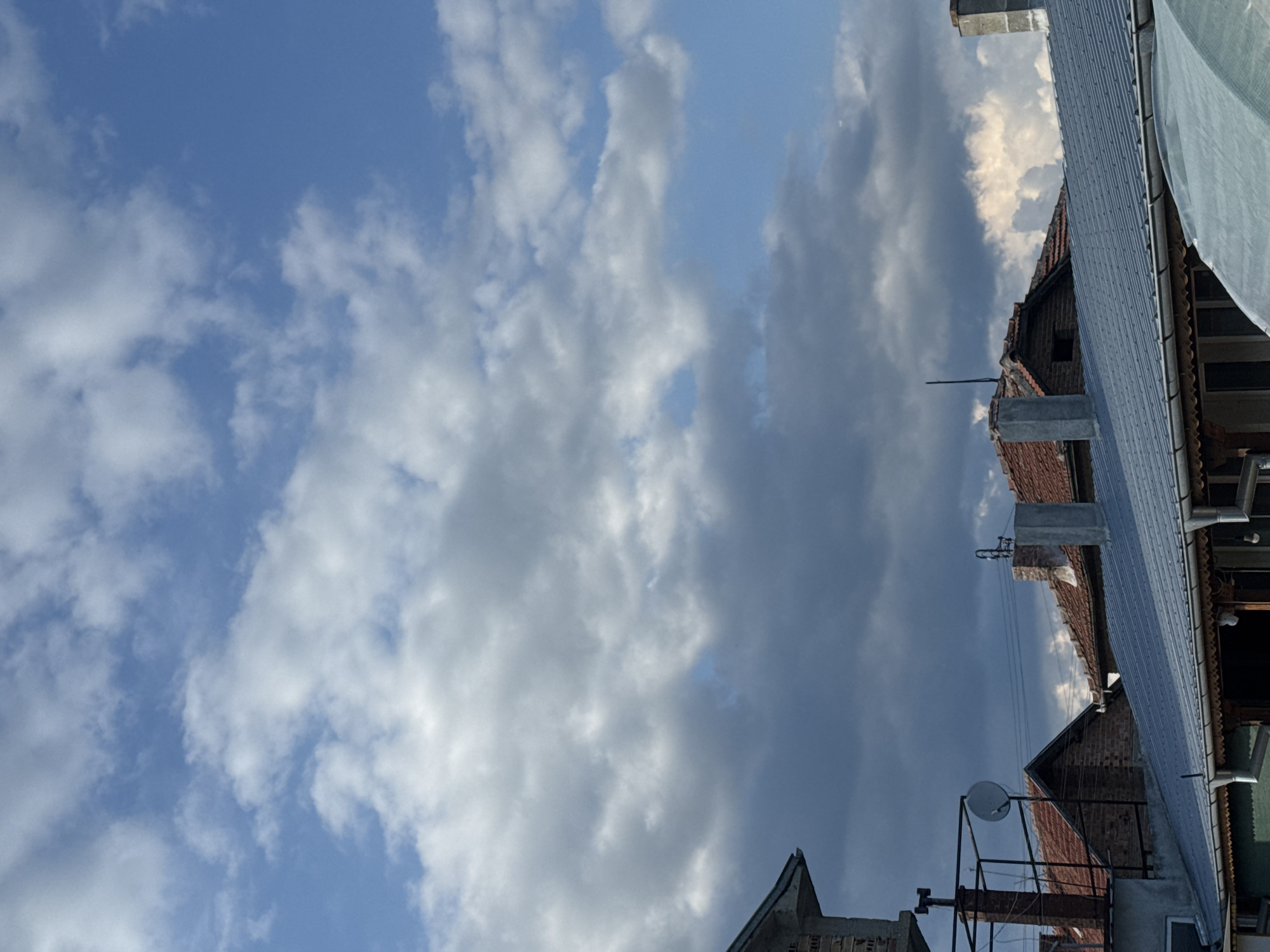 The image shows a view upwards towards the sky, partially obscured by buildings and infrastructure. The sky is blue with scattered white and grey clouds. The buildings appear to be residential, with red tiled roofs, chimneys, and metal roofing. A satellite dish is visible, mounted on a metal frame. There are also pipes, wires, and other attachments on the buildings. The photo location is in Sarnevo, Bulgaria.