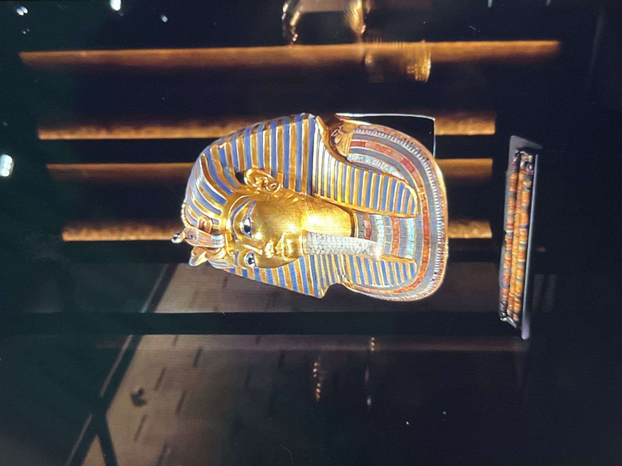The image shows a golden mask, possibly Tutankhamun's, displayed on a dark surface. The mask has blue and gold stripes, with a gold face and patterned details. Next to the mask is a narrow, rectangular object with similar decorative patterns. The background consists of horizontally aligned, illuminated bars. The image's setting appears to be a museum or exhibition. There is no indication of the location being in Tinaquillo, Venezuela.