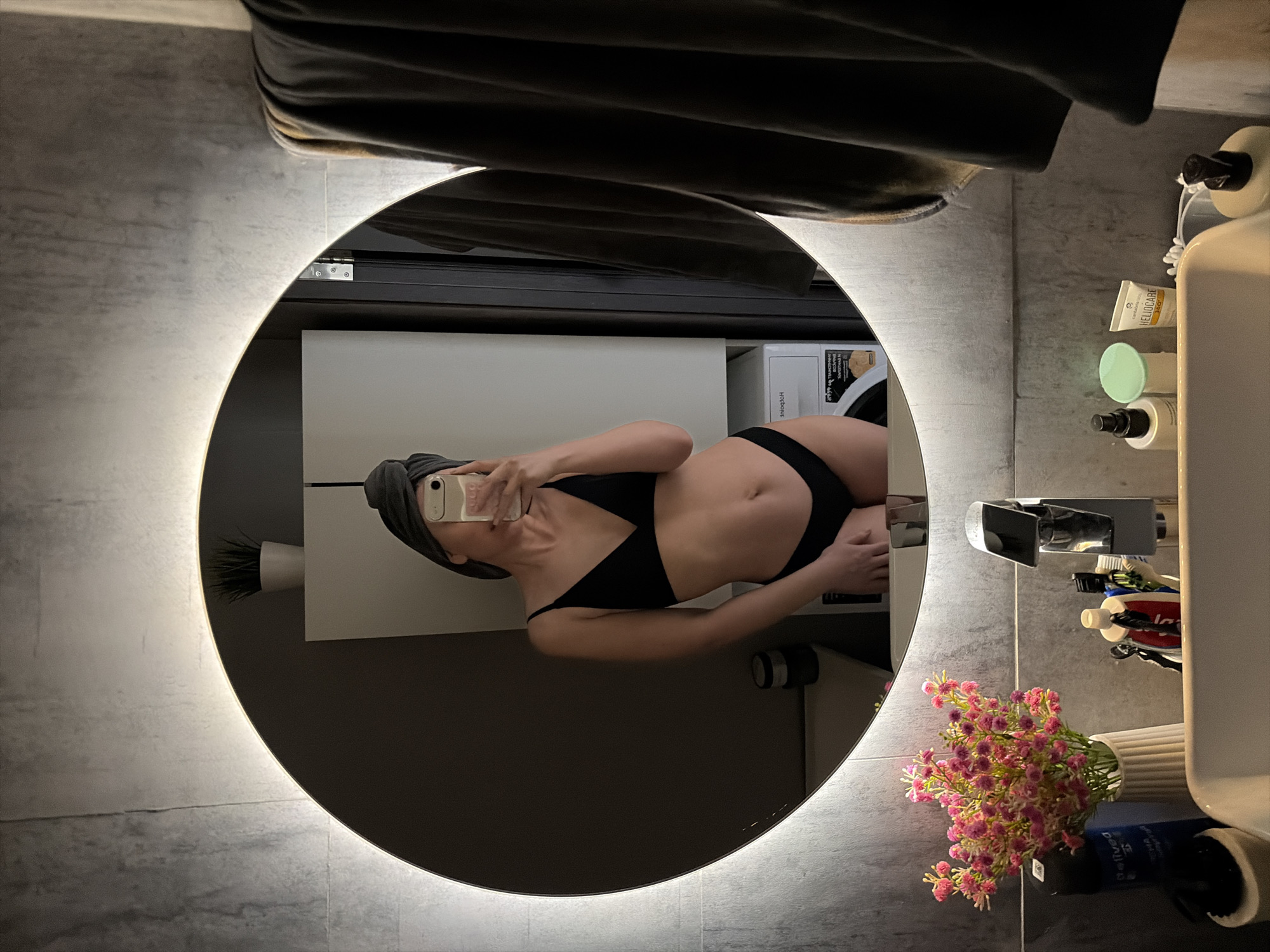 A mirror selfie shows a woman in a black bikini and a towel wrapped around her head, holding a cell phone to take the picture. The reflection is in a round, illuminated mirror in a bathroom. The bathroom features grey tiling, a white cabinet, toiletries, and a vase of artificial flowers near the sink. The image offers no clues about being located in Chaikovsky, Russia.