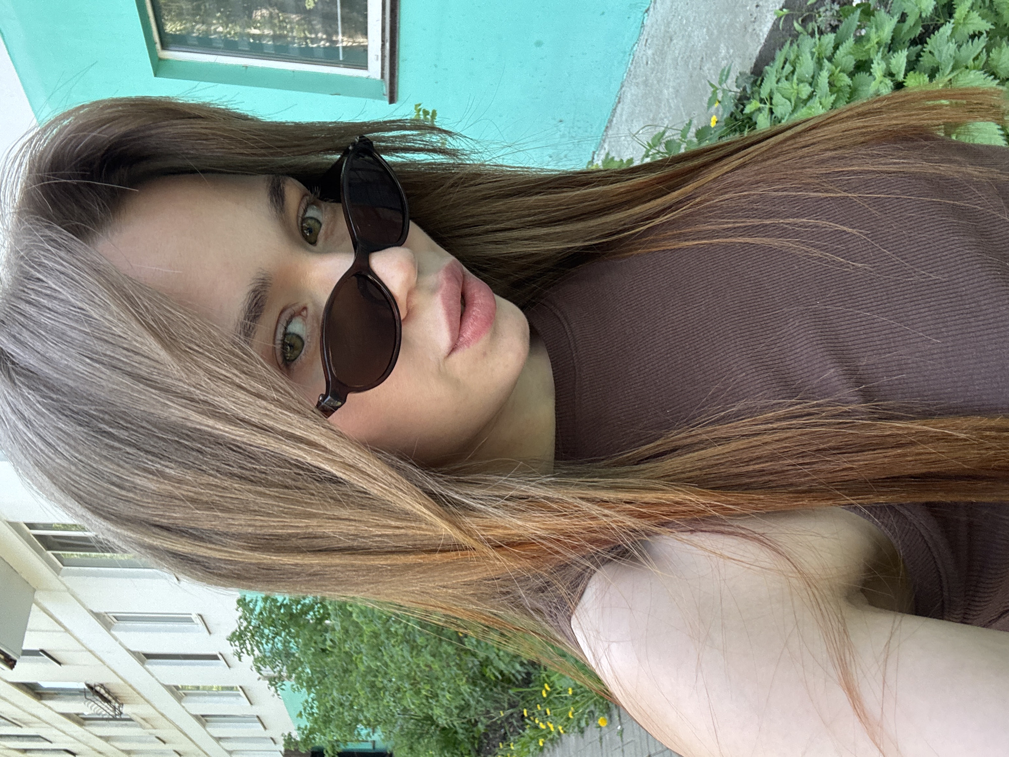 A woman with light brown hair and wearing sunglasses is shown in a vertically oriented medium shot. She is wearing a brown top. The background includes a green building with a white-framed window and a sidewalk with green plants. A white building with windows and green trees is in the lower left corner of the frame. The location is likely Chaikovsky, Russia.