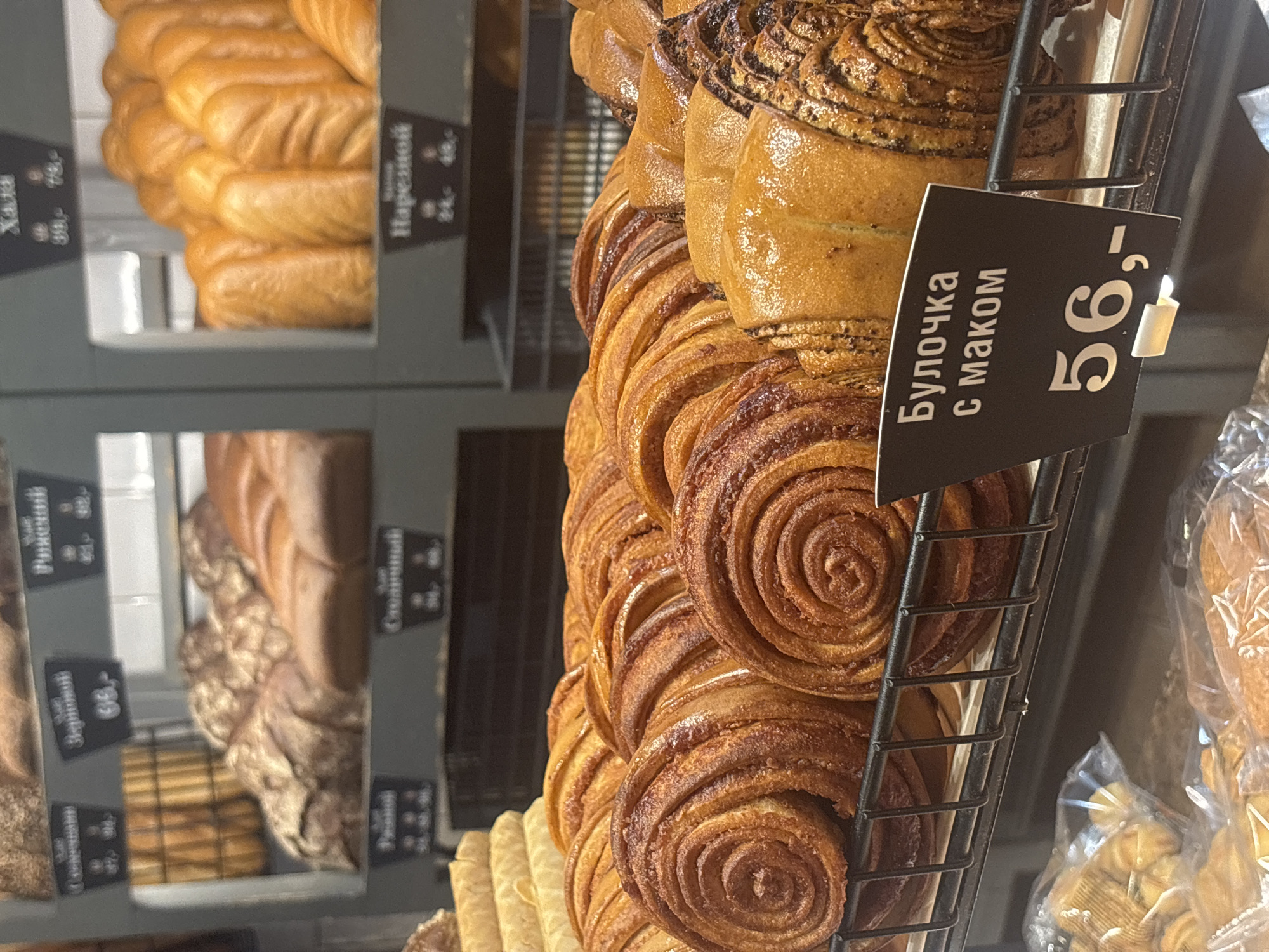 The image shows a close-up of baked goods displayed on shelves in a bakery or food store. In the foreground, there are several spiral-shaped pastries with a cinnamon or sugar filling, appearing golden brown and slightly glazed. A price tag in Russian, reading "Булочка с маком" (Bun with poppy seeds) and "56,-", is placed in front of these pastries. Behind them, on the same shelf, are more pastries, some with a visible poppy seed filling and others with a plain golden-brown crust. The shelves are made of metal wire. In the background, other baked goods such as bread loaves and rolls are visible on higher shelves, some with Russian labels. The overall environment suggests a retail setting for fresh bread and pastries. The image does not provide specific information about the location within Saint Petersburg, Russia, but it depicts a typical bakery display. There are no people visible in the frame.