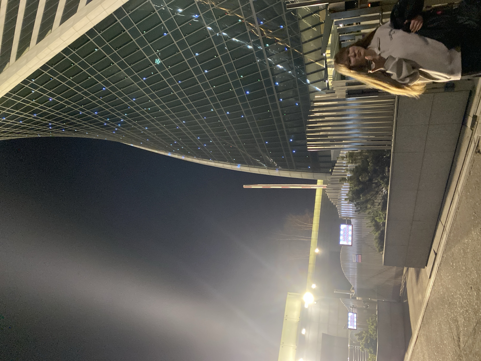 The image depicts a nighttime urban scene in Khimki, Russia. In the upper right, a person with long blonde hair, wearing a light-colored sweatshirt, is visible. A building with a curved, grid-patterned facade illuminated with small blue lights dominates the upper left. Below, the night sky is visible. To the right, there is street lighting and signage in Russian. The scene includes greenery and elements of modern architecture.