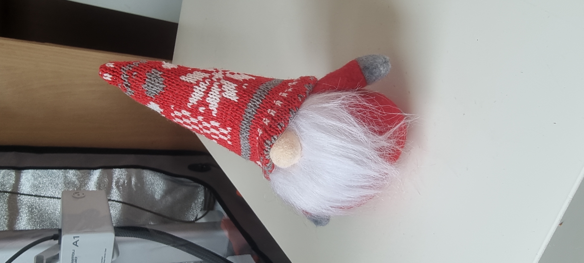 A knitted gnome with a red and white snowflake patterned hat, white beard, and red body sits on a white surface. The gnome is positioned slightly to the right of the image center. In the background, to the left, is a white electrical adapter with the label "A1" and a dark grey fabric bag. To the right of the gnome, a portion of a wooden surface is visible. The scene appears to be indoors, likely on a table. The location is not identifiable as being in Naaldwijk, Netherlands.