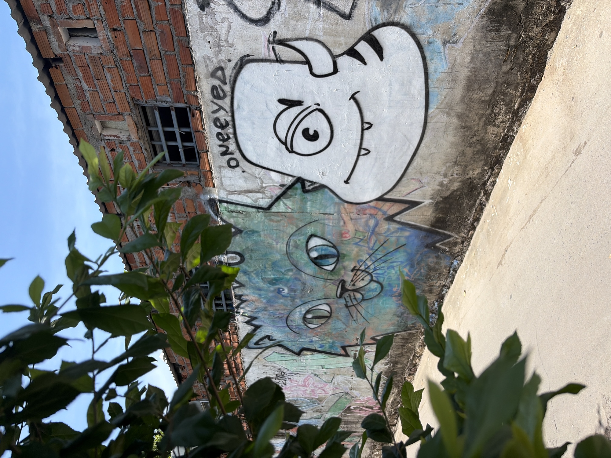 The image captures a section of a wall covered in graffiti, located in the city of Bac Nha Trang Ward, Vietnam. The wall itself is concrete and appears weathered, with evidence of water stains and dirt. Above the wall, a building constructed of red bricks is visible, with some windows covered by metal bars. The roof is made of corrugated metal. In the foreground, a pathway made of concrete runs along the base of the wall. Lush green foliage from a tree partially obscures the view of the wall and the building.
The graffiti on the wall features two distinct characters. On the left, a stylized cat with large blue eyes and a playful expression is painted in shades of blue and green. To the right of the cat, a white, cartoonish monster with a single eye, sharp teeth, and striped ears is depicted. The word "oneeyed" is spray-painted above the monster. Other, less distinct graffiti tags and drawings are also present on the wall. The sky is clear and blue. No people are visible in the image.