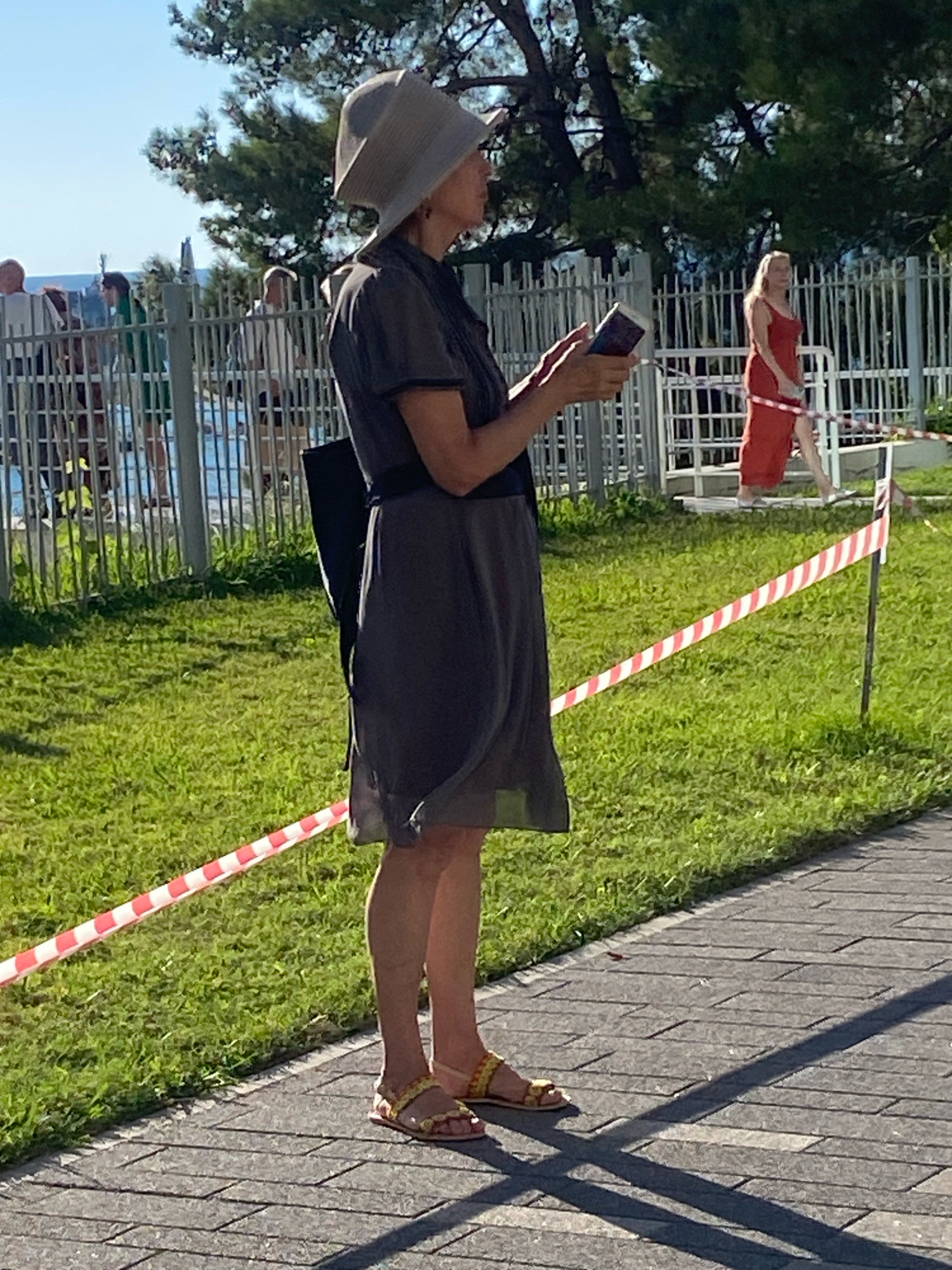 The image depicts a woman standing on a paved pathway, looking at a mobile phone. She is wearing a wide-brimmed hat and a dark dress, with yellow sandals on her feet. Behind her, a red-dressed woman is visible near a metal fence. The environment appears to be outdoors, with a grassy area and trees in the background, and a clear blue sky above. The scene is likely set in Sochi, Russia, given the presence of a beach or seaside location hinted at by the background, and the general appearance of the foliage and fencing.