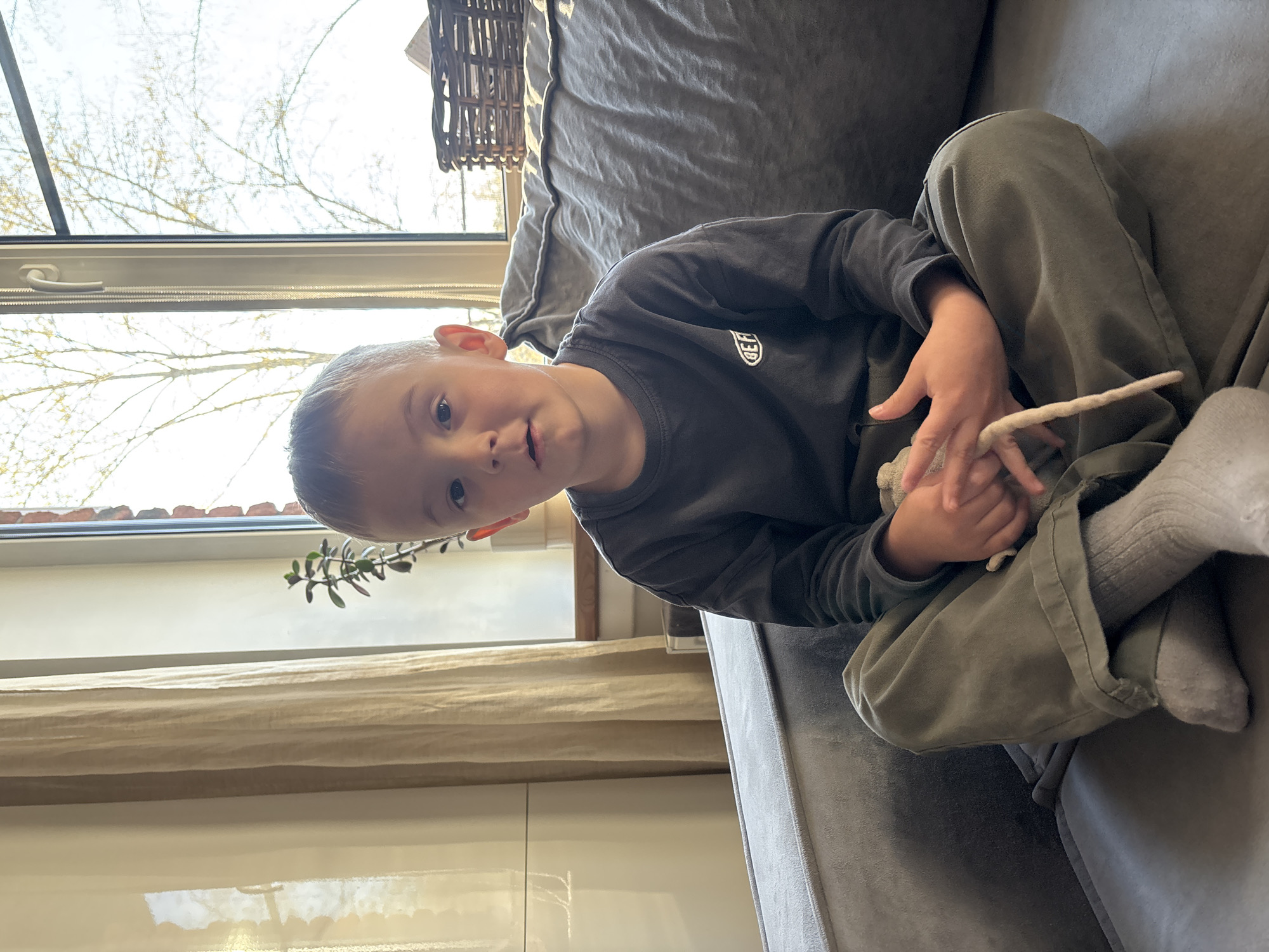 A young boy is sitting cross-legged on a gray couch. He is wearing a dark gray long-sleeved shirt, olive green pants, and gray socks. He is holding a long, light-colored object in his hands. In the background, there is a window with a view of bare trees and buildings. The interior space also contains a woven basket on top of the couch, neutral-toned curtains, a small potted plant, and a light-colored, shiny cabinet. The location is Chaikovsky, Russia.