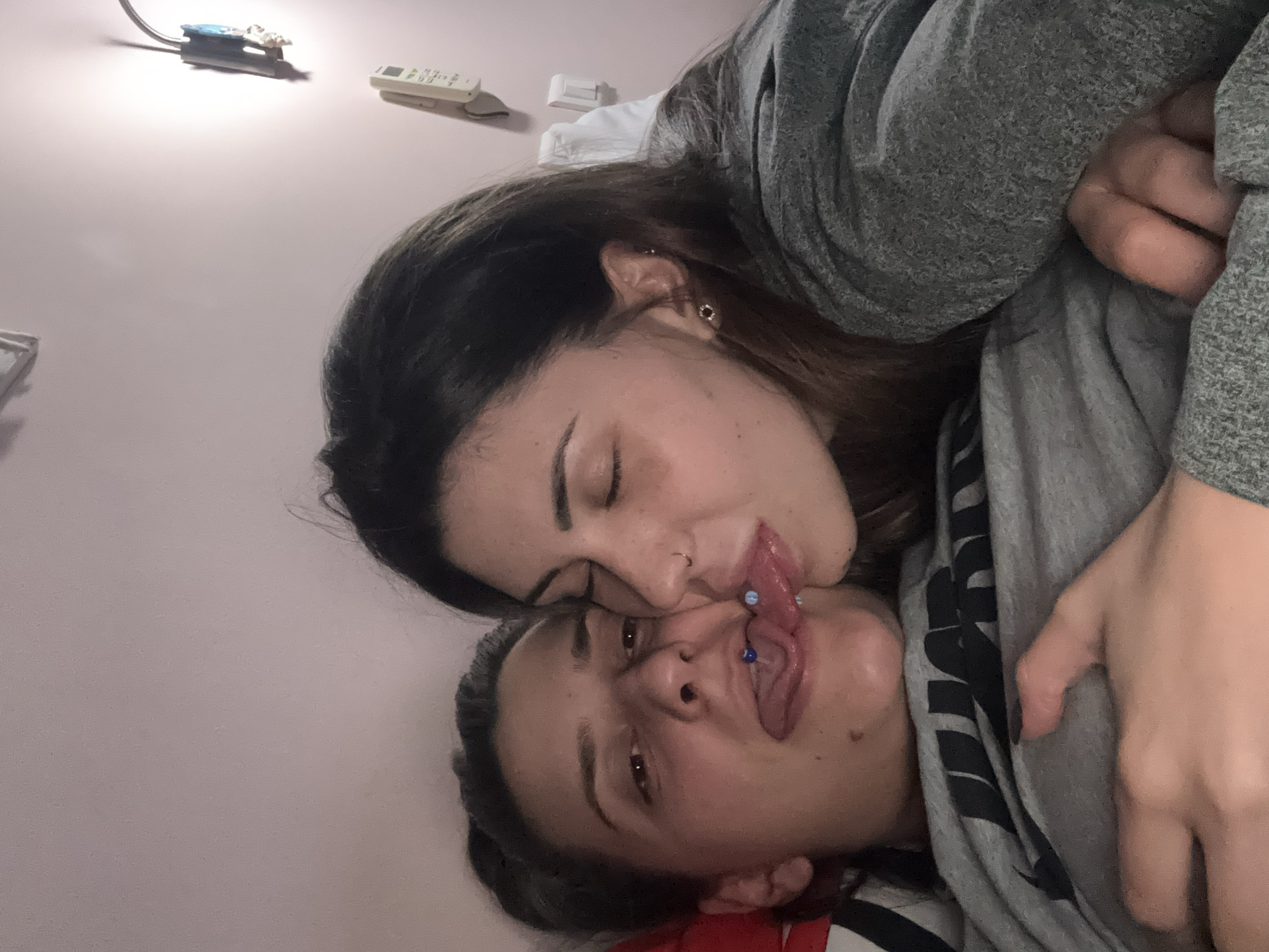Two women are intimately entangled, their faces close. One woman lies supine with her tongue extended, revealing blue piercings. The other woman leans over her, her tongue touching the exposed tongue. Both are wearing gray clothing. The environment appears to be an interior setting, likely a bedroom, suggested by the presence of a lamp, remote control, and a wall. There is no discernible information to place the scene in Sofia, Bulgaria.