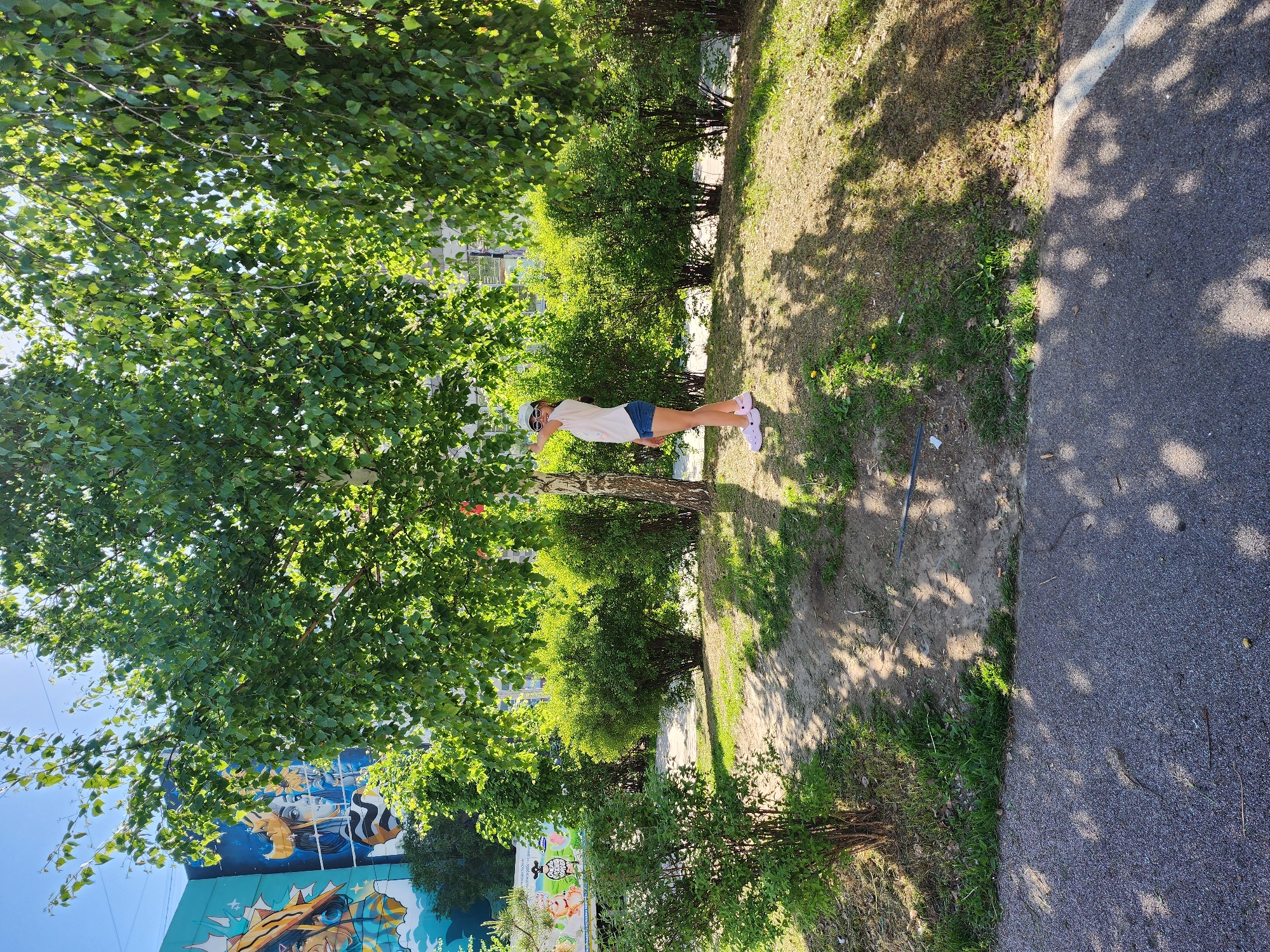 The image shows a park-like setting in Kazan, Russia, based on details like the architectural style and possibly Cyrillic text on buildings. A woman wearing shorts and light clothing stands near a tree. There is foliage including trees and bushes, and a paved path adjacent to the grassy area. There is a wall or building with a mural visible in the background.