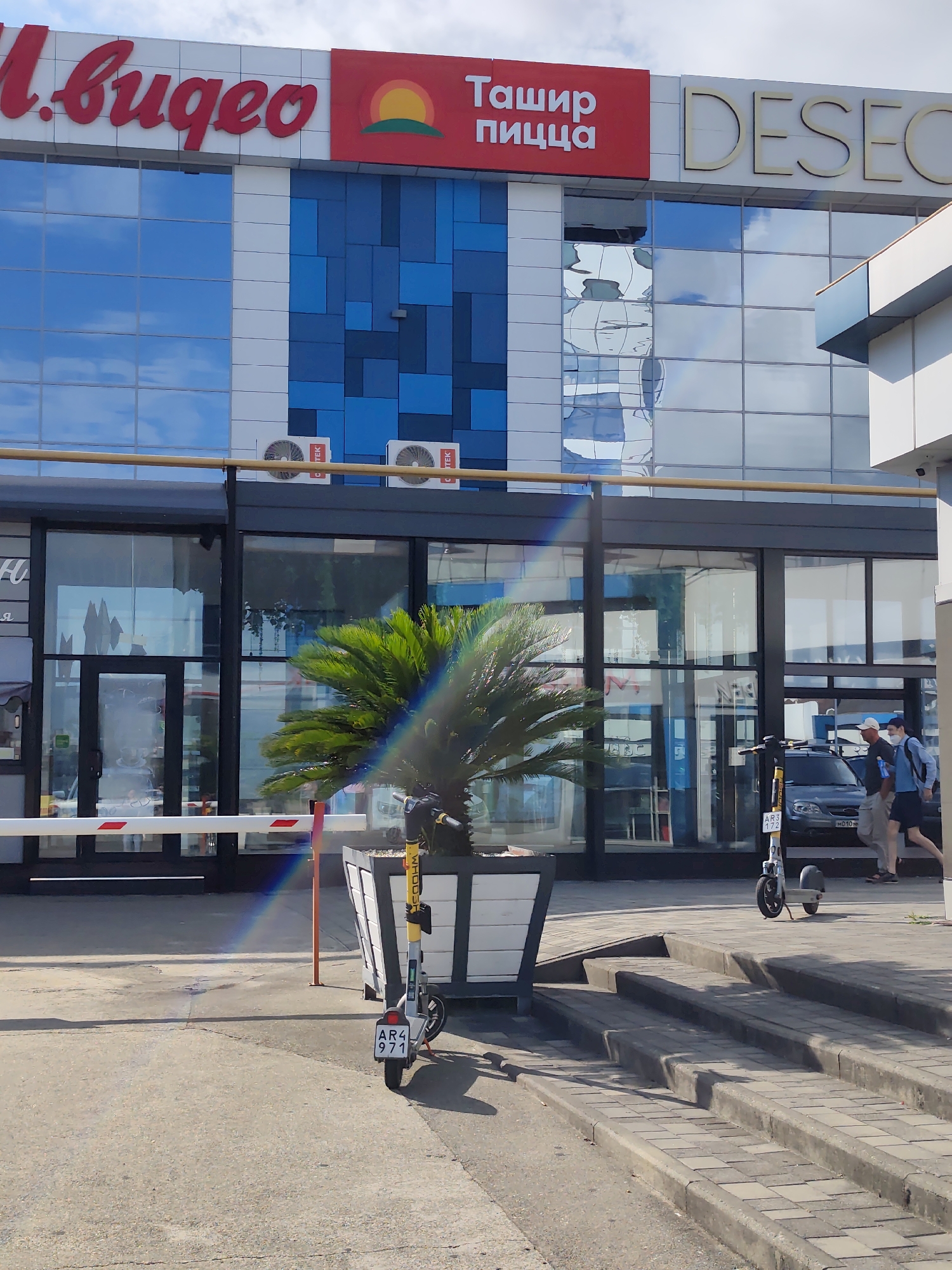 In Sochi, Russia, a commercial building features prominent signage, including a red sign with the text "Ташир пицца" (Tashir Pizza) and a logo of a sun over green hills. Adjacent to this is a gold-lettered sign that reads "DESECO". The building has a facade of blue and white tinted windows reflecting the sky. Two outdoor air conditioning units are mounted on the exterior wall. In front of the building, a white barrier gate with red and white reflective strips is partially lowered. A planter box contains a palm-like plant. A white and yellow electric scooter, license plate "AR4 971", is parked next to the planter. Two men are walking past the building; one, wearing a light-colored cap and a mask, is walking to the left, and another, wearing a blue shirt and shorts, is walking to the right. Further to the right, two more people are walking, one with a backpack and the other with a blue shirt. A grey car is parked behind the scooter. The ground in front of the building is paved with concrete and pavers, with a set of concrete stairs leading up to a sidewalk. A rainbow-like light refraction is visible across the scene.