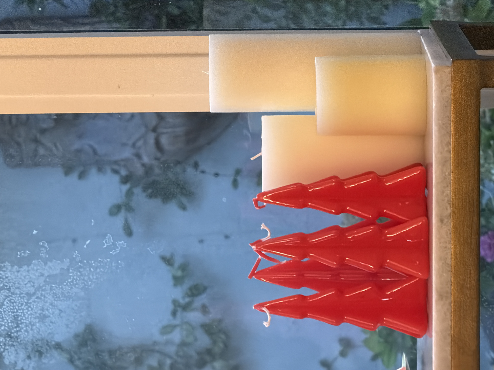 A horizontal close-up shot captures a festive display of red Christmas tree-shaped candles and white pillar candles on a marble shelf in San Marino, California. The red candles, arranged in a cluster of five, have white wicks extending from their pointed tops. To their right, three white pillar candles of varying heights are placed. The background, visible through a window, is blurred and displays a blue-toned scene with hints of greenery and what appears to be a stucco statue. The overall impression is one of holiday cheer, set against a backdrop of what seems to be a residential or commercial interior.