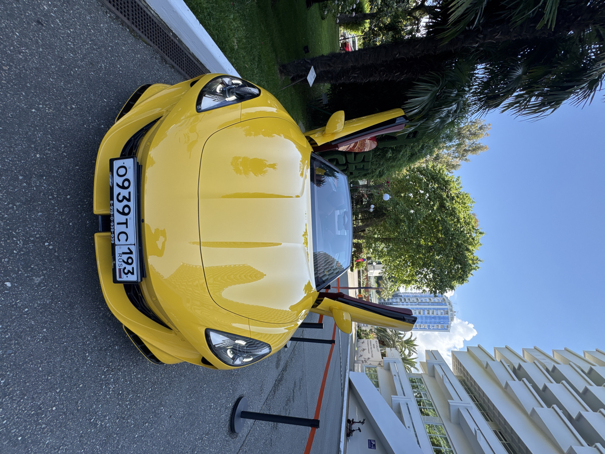 A bright yellow Ferrari sports car with its scissor doors open is parked on an asphalt road in front of a white modern building. The car has a Russian license plate that reads "0939TC 193 RUS." To the left of the car, there is a green lawn with palm trees and other greenery. In the background, there is a tall blue building and a large white building with balconies. The sky is clear and blue. The scene appears to be in a resort area. The location is likely Sochi, Russia, given the presence of palm trees and a resort-style building, and the license plate format.