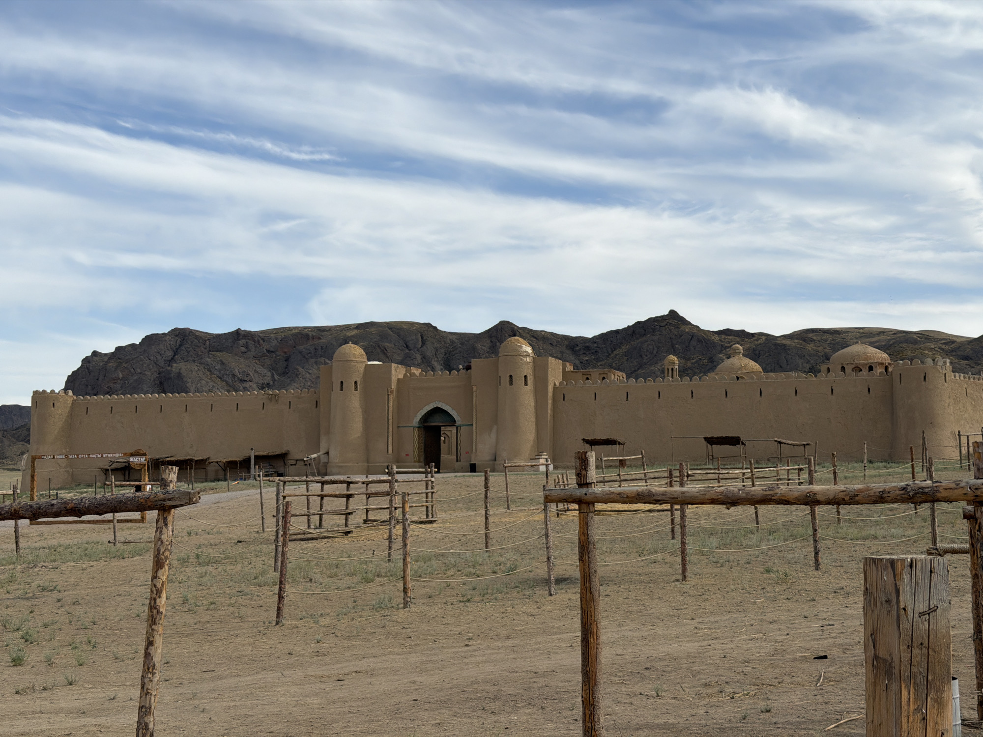 The image depicts an exterior view of a historical fort in Qonaev, Kazakhstan, under a partly cloudy sky. The fort, constructed from earth-toned materials, features crenellated walls, cylindrical towers, and domed structures. A large arched gateway serves as the main entrance, with a sign above it in a script that appears to be Cyrillic or a similar alphabet. In the foreground, a series of wooden fences and posts are arranged, suggesting an enclosure or pen, possibly for livestock or as part of a historical reenactment setting. The ground is dry and dusty, with sparse vegetation. The overall scene evokes a sense of history and perhaps a movie set or historical park, given the weathered appearance and the rustic fencing. The light suggests it is daytime, likely afternoon, with soft shadows. There are no people visible in the frame, and the environment appears serene and quiet. The mountains in the background add to the arid, rugged landscape characteristic of the region.