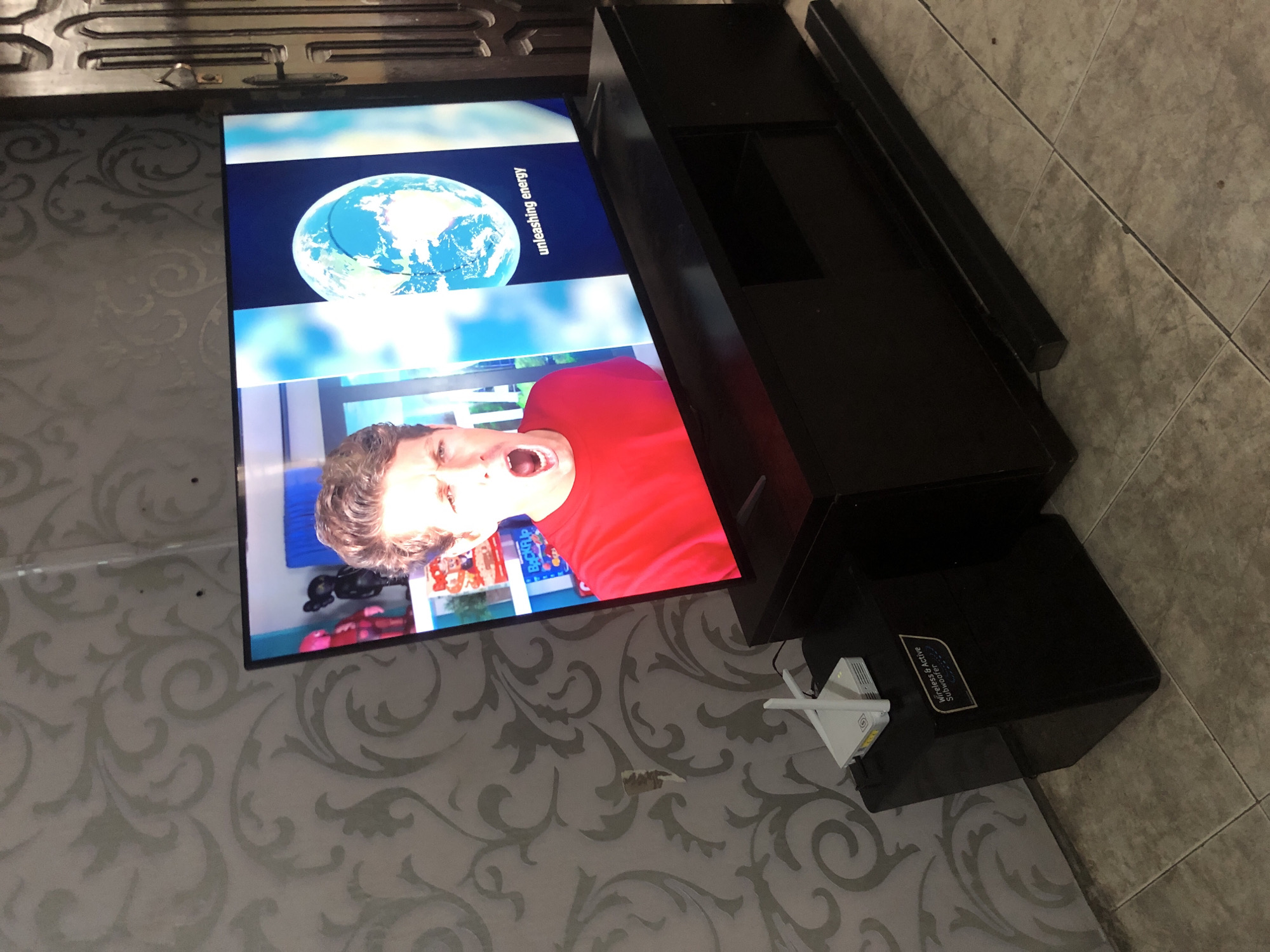 A television set is centered in the frame, displaying a man with his mouth open in a shout, wearing a red shirt, and an image of the Earth with text that reads "unleashing energy." The television is placed on a dark brown entertainment stand. To the left of the television, on a black subwoofer, is a white wireless router. The background features a grey patterned wallpaper. The scene appears to be indoors, likely in a living room, with a tiled floor. A dark wooden door is visible on the right edge of the frame. The image was taken in Ado, Nigeria.