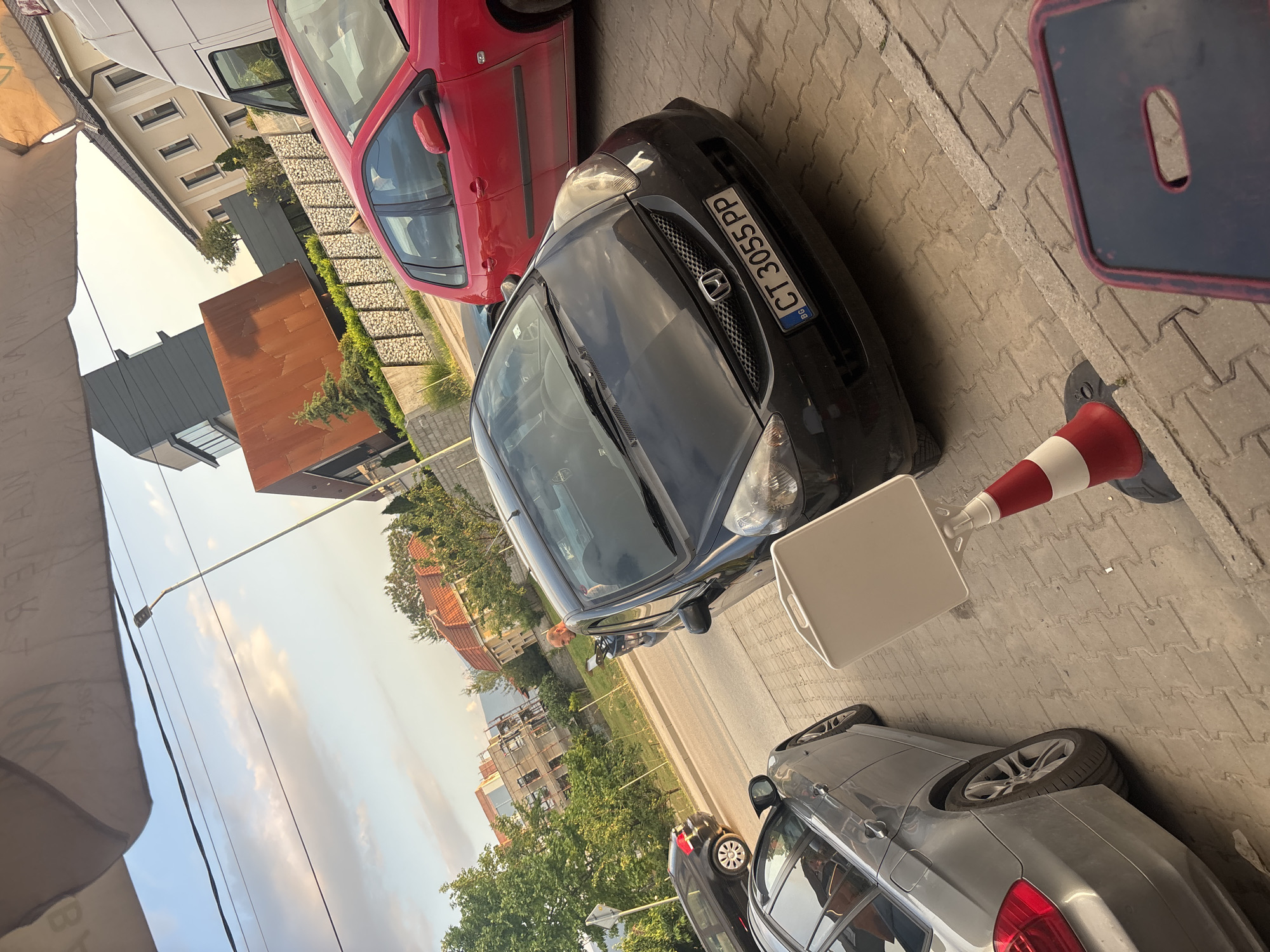 The image shows a street scene in Plovdiv, Bulgaria, with several parked cars. A black Honda with a Bulgarian license plate (CT 3055 PP) is parked alongside a red car and a silver BMW. A red and white traffic cone is positioned near the Honda. A small, white folding table is open near the BMW. Buildings are visible in the background. The sky is overcast.