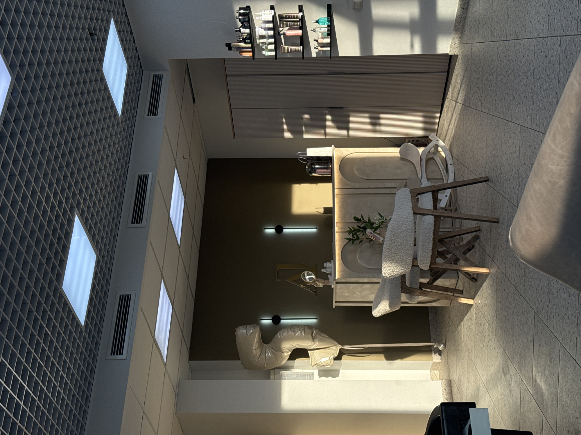 The image shows the interior of a room in an unknown location. The space is well-lit with a combination of natural light and square, recessed ceiling lights. The floor is tiled with grey speckled tiles. A light beige vanity with a rounded basin and decorative items stands in the center of the room, beside a wooden chair covered in soft white material. On the left side of the image, there is a patterned ceiling design with several rectangular air conditioning vents. Above the vanity, there are shelves displaying bottles and containers, possibly beauty or personal care products. The walls are a combination of light beige and a darker shade of brown. Some linear lighting fixtures are present on the darker wall. A beige inflated cushion sits near a lower wall. A partial view of a black chair and a light beige colored object is visible in the foreground. The room gives the impression of a modern, minimal spa or salon.