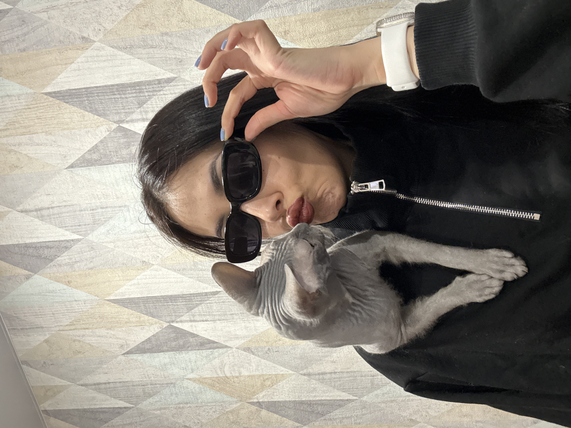 A young adult female is shown, wearing a black zip-up sweatshirt, black sunglasses, and a white watch. She is holding the sunglasses on her face with her right hand and pouting her lips. A hairless cat is resting in the front of her jacket. The background is a geometric wallpaper of triangles with neutral colors. No information is available to indicate a location in the city of Votkinsk, Russia.