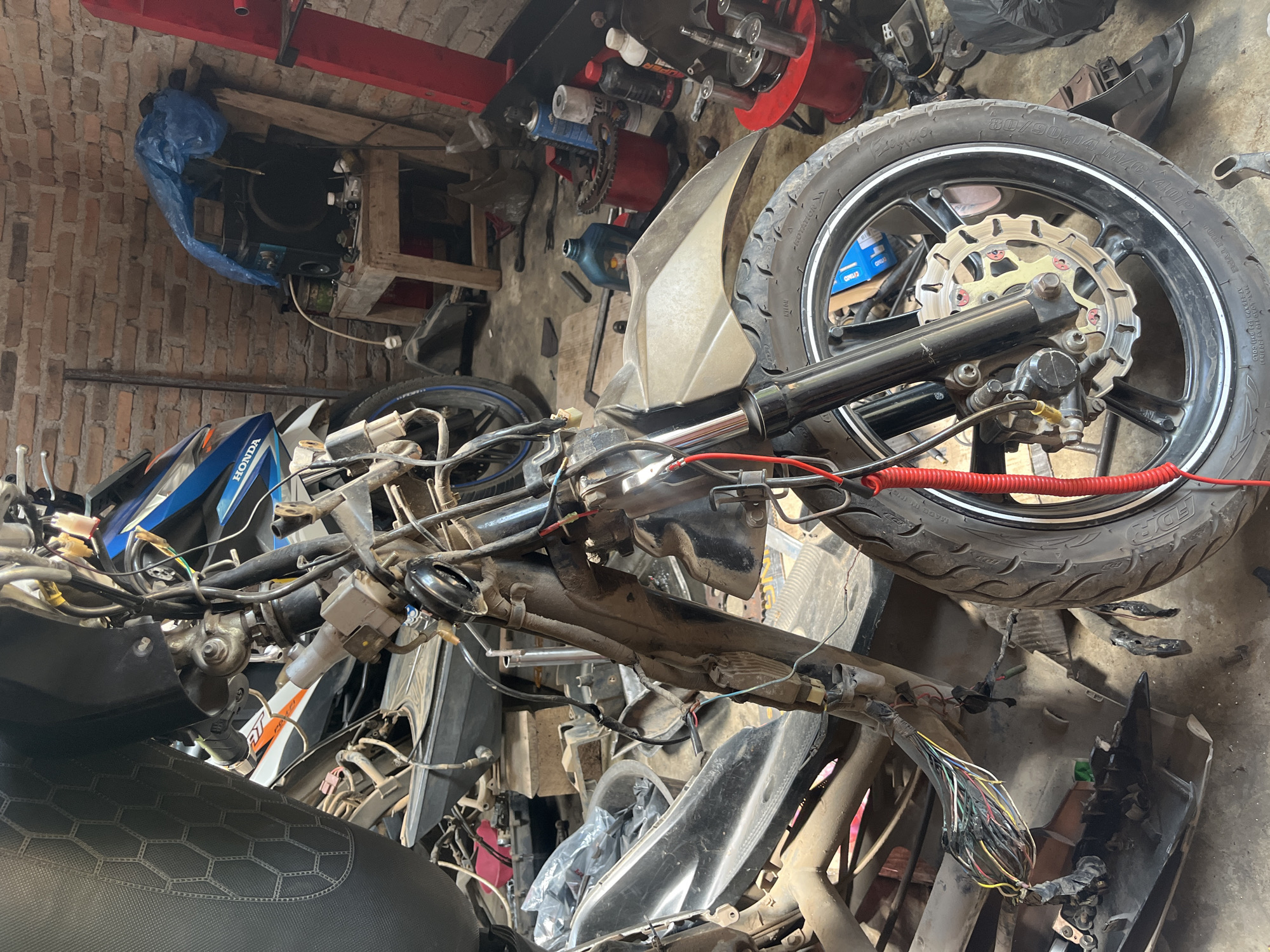In a dimly lit garage in Kebumen, Indonesia, a partially disassembled motorcycle lies on its side. The front wheel and forks are prominent, with exposed wiring and internal components visible. The motorcycle's seat, handlebars, and frame are also visible, along with various tools and spare parts scattered around the workspace. The background reveals a brick wall and shelves filled with items, suggesting a functional repair shop.