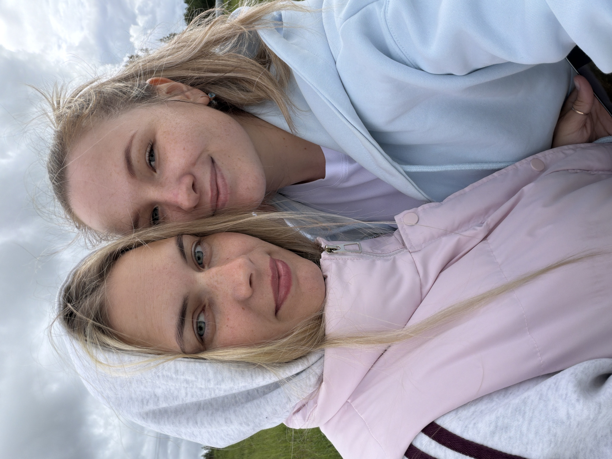 Two young women are posing for a selfie outdoors. The woman on top wears a light blue hoodie over a white top. The woman below wears a grey hoodie and a light pink jacket. The background shows a cloudy sky and some green grass. The location is Ольховка, Russia.
