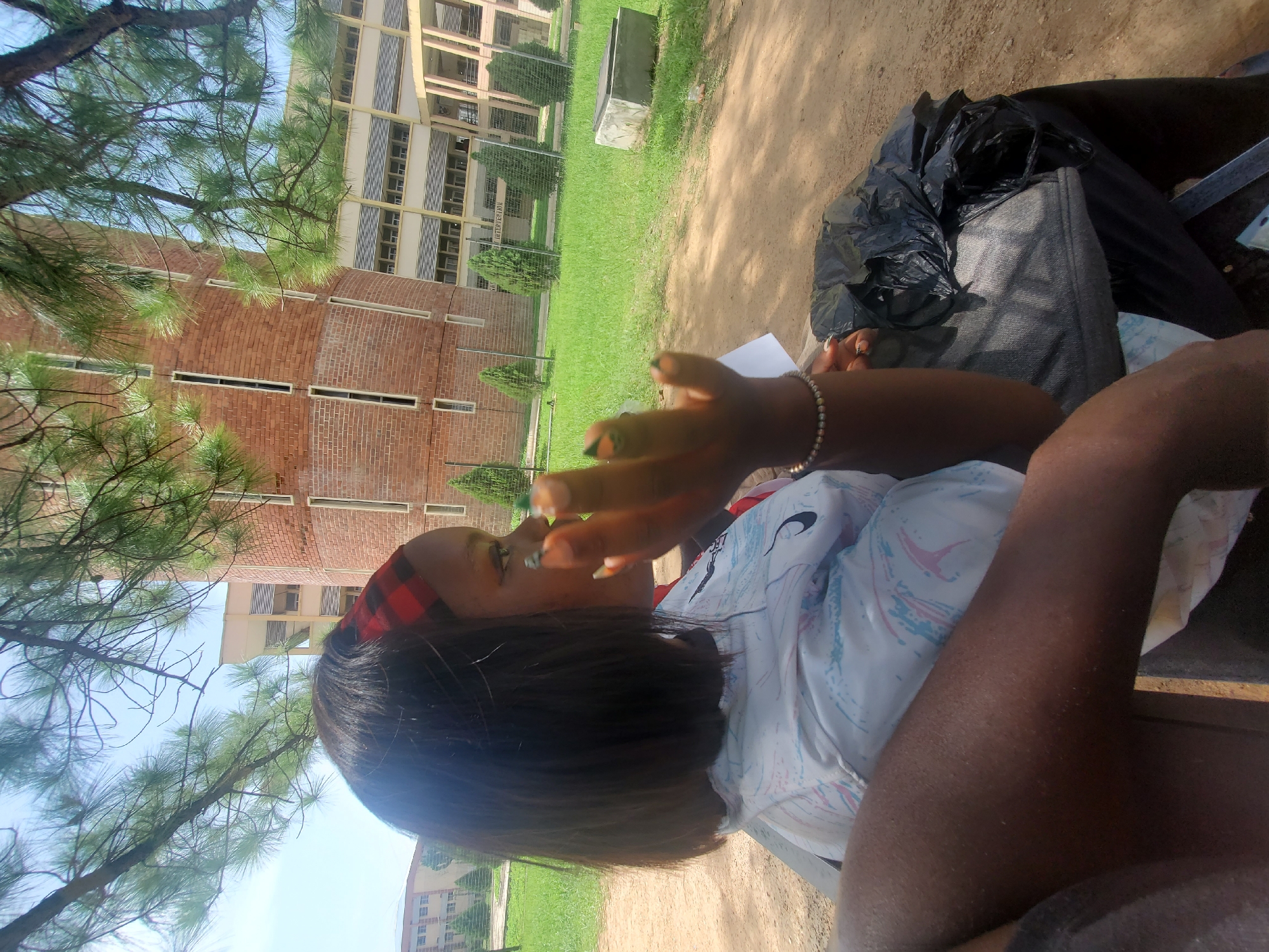 The image shows a young woman sitting outdoors, possibly on a bench or in a chair, in what appears to be a university campus environment. She is wearing a white t-shirt with a pattern of pink and blue lines. A red and black checkered headband is on her head, and her hair is dark and short. She is holding a piece of paper and appears to be looking at it, with her right hand raised, suggesting she might be reading or writing. Her nails are painted with dark green polish.
In the background, a modern brick building with multiple stories and numerous windows is visible. The building has a light-colored facade and vertical window louvers. Adjacent to the building is a well-maintained green lawn, bordered by a metal fence and some small, manicured evergreen trees. Pine trees with their distinctive needles are in the foreground, partially obscuring the view. The ground around the woman is dirt and sand. A black plastic bag is placed next to her.
The lighting suggests it is daytime, likely a sunny day, given the bright sky and shadows. The presence of lush green grass and trees indicates a warm climate. The overall setting appears to be a casual outdoor area conducive to studying or relaxing. No specific text is clearly legible in the image.