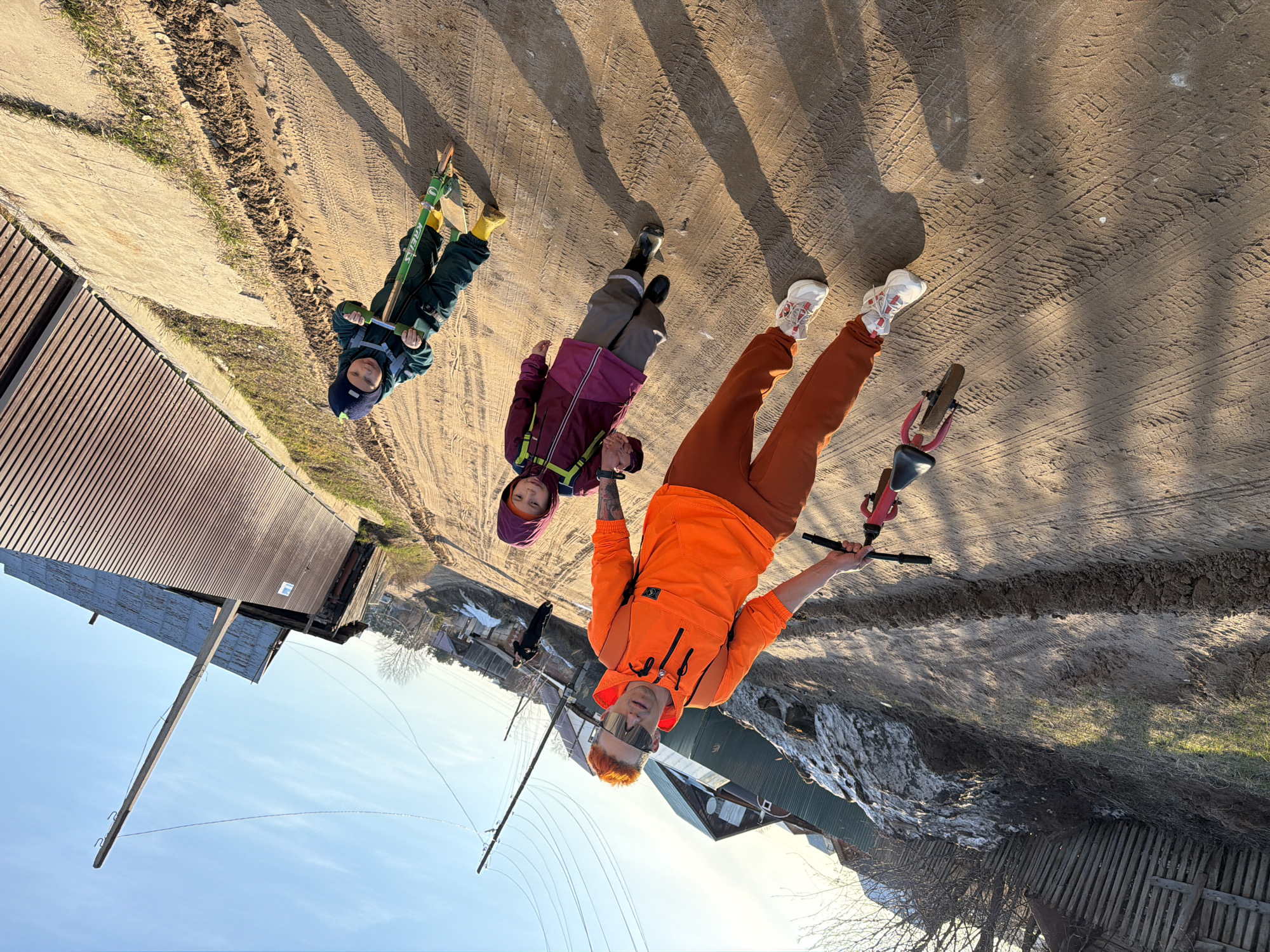 The image shows three children and an adult in an outdoor setting on a dirt road or path, likely a rural or suburban area. The image seems to be taken in the city of Chaikovsky, Russia.
The first child on the left is wearing a dark blue hat and green snowsuit, riding a scooter with green and yellow colors. The second child, standing next to the first, is wearing a purple jacket with grey pants and black boots. The adult in the center of the image is wearing an orange sweatshirt and pants with white sneakers with red accents. The adult is holding a small red and black bicycle.
The background includes residential structures with wooden exteriors, some with snow on the roofs, and bare trees. The dirt road or path shows tire tracks. The sky is clear and blue.
