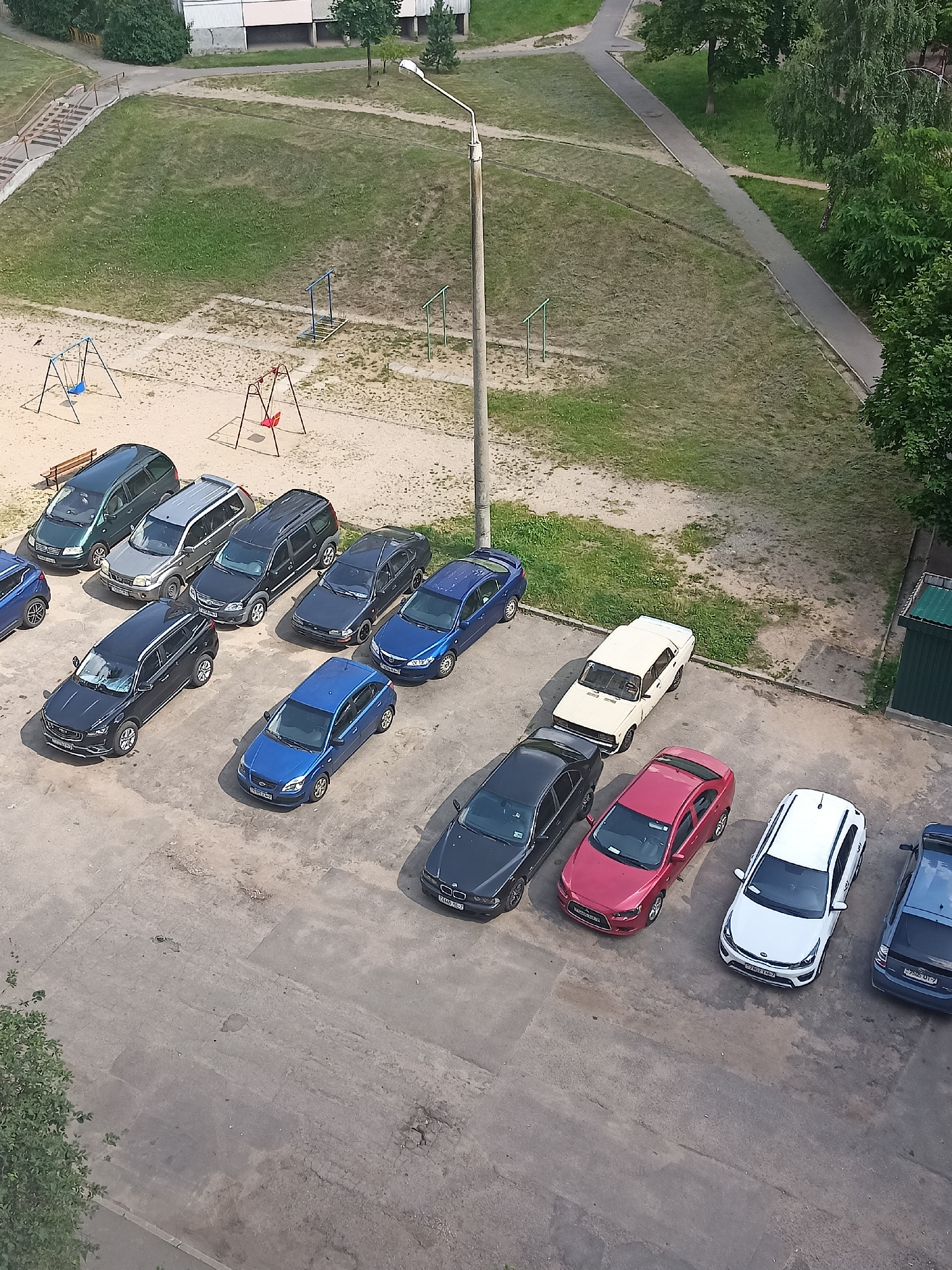 This is an aerial photograph of an outdoor parking lot in the city of Minsk, Belarus. Multiple cars are parked in rows on a paved surface. In the background, a playground with swings and jungle gyms is visible, adjacent to a grassy area and a building. A lamppost stands near the playground. The weather appears to be fair and sunny. No people are visible in the scene.