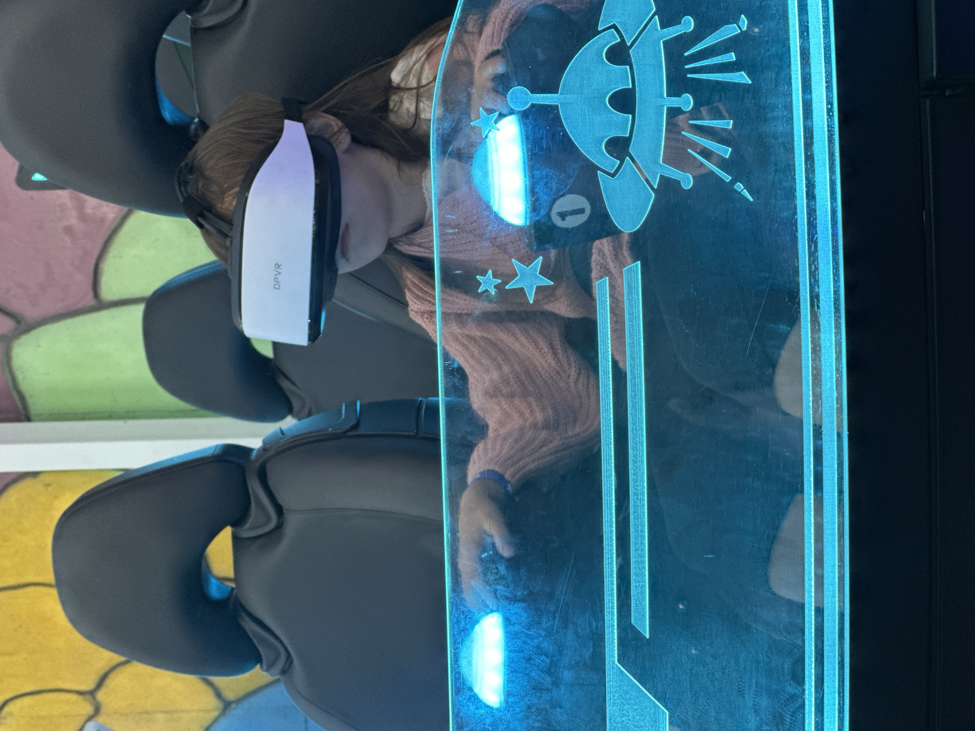 A young girl wearing a white VR headset with "DPVR" printed on it is seated in a black, futuristic-looking chair. She is looking to her right and wearing a light pink knitted sweater. In front of her is a transparent blue panel with illuminated graphics of stars, a flying saucer, and the number "1". The background consists of a colorful, abstract mural with yellow, green, and pink shapes. This image was captured in Vityazevo, Russia.