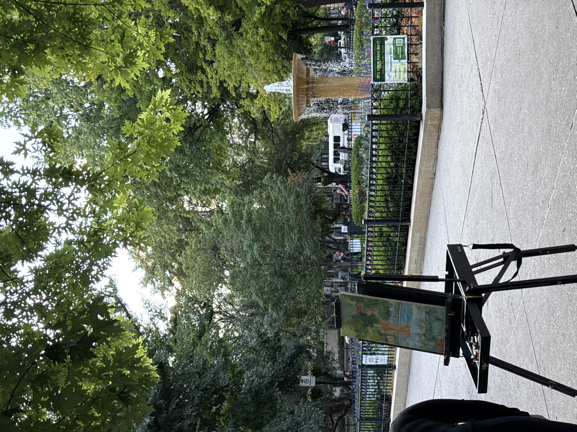 This image depicts a park scene in Chicago, Illinois, United States of America, with an easel and a painting in the foreground. The painting appears to be a landscape, possibly of the park itself, with a fountain visible in the background. The easel is set up on a paved walkway, and a person is partially visible to the left, standing behind the easel.
In the middle ground, a black metal fence encloses a garden area with blooming flowers. Beyond the fence, a fountain with water spraying upwards is the central focus. A white bus or shuttle is parked behind the fountain, and several people are visible in the background, some standing and others walking. Trees with lush green foliage dominate the background, suggesting a warm season.
A sign with text is visible to the right of the fountain, indicating "IT'S YOUR PARK DAY!" and mentioning dates and times, implying an organized event. Another sign with a circular symbol is visible on a pole behind the fence to the left. The overall atmosphere is serene, with the sounds of the fountain likely contributing to the ambiance. The lighting suggests it is daytime, possibly late afternoon given the angle of the sun.