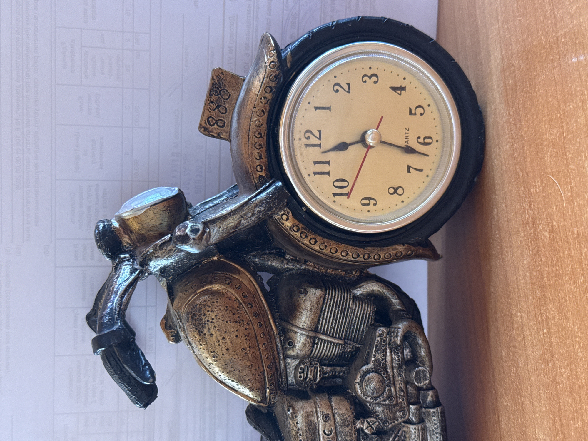 A close-up, eye-level, medium shot captures a decorative clock shaped like a motorcycle. The clock face, a creamy yellow with black numerals, is partially obscured by the motorcycle's wheel. The hour hand points to the 11, and the minute hand points to the 6, indicating 11:30. A thin red second hand is visible. The motorcycle, constructed from a bronze-colored material with metallic accents, features intricate detailing on its engine and bodywork. A small, rectangular plaque with the number "888" is affixed to the top of the motorcycle. The background consists of a white paper with rows of text. The object rests on a light brown wooden surface.
