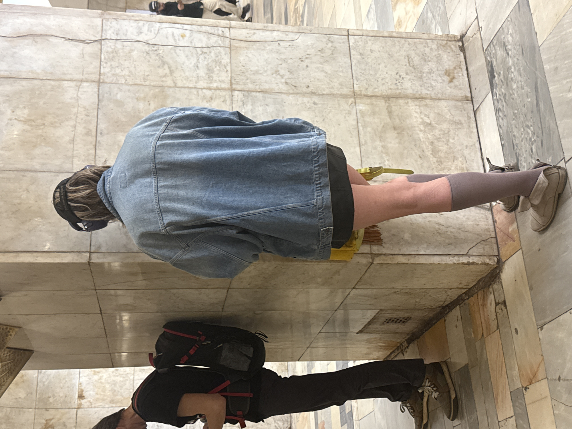 A medium shot, with the camera directly overhead, captures two individuals on a marble floor. The first individual is kneeling and appears to be sweeping with a hand broom. The person is clad in a denim jacket and dark shorts, and they are wearing knee-high gray socks and tan shoes. A pair of headphones rests atop their head. Beside them, a yellow and green broom is partially visible. The second individual is seen standing, wearing a black shirt, black pants, and tan shoes. A black and red backpack rests on their back. Both are located inside the Moscow Cathedral Mosque, recognizable from the patterned marble tiles and architectural details.