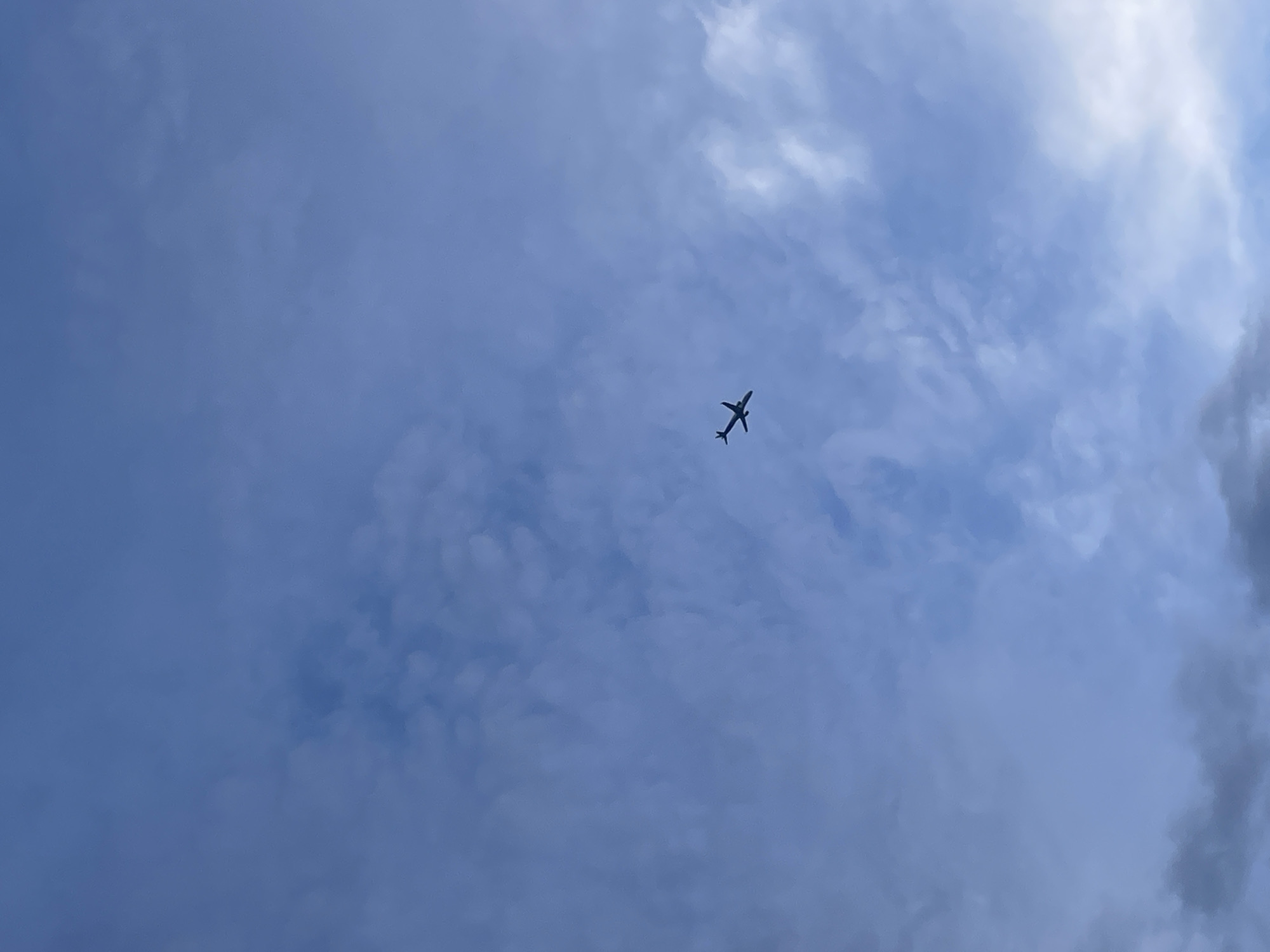 The image shows a wide shot of a blue sky with scattered clouds. A small airplane is visible in the center of the frame, silhouetted against the sky. The airplane is a dark color and appears to be in flight. The clouds are wispy and white, and they are spread throughout the sky. The overall impression is one of a clear day with a single aircraft in the distance. This image was taken in Budva, Montenegro.
