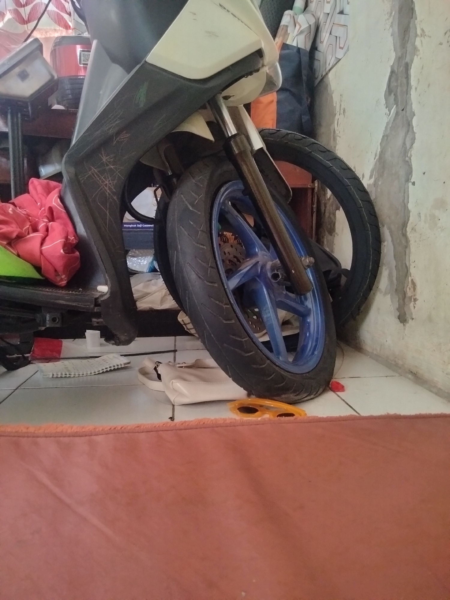 The image shows a motorcycle parked indoors, leaning against a textured, off-white wall. The front tire of the motorcycle, with a blue rim, is prominent in the foreground. The motorcycle's bodywork has visible scratches. To the left of the motorcycle, a red cloth is draped over something obscured from view. On the tiled floor in front of the motorcycle, a white handbag, a spiral notebook, and a pair of yellow sunglasses are visible. The scene suggests an indoor storage area, possibly a home, in Cariu, Indonesia.