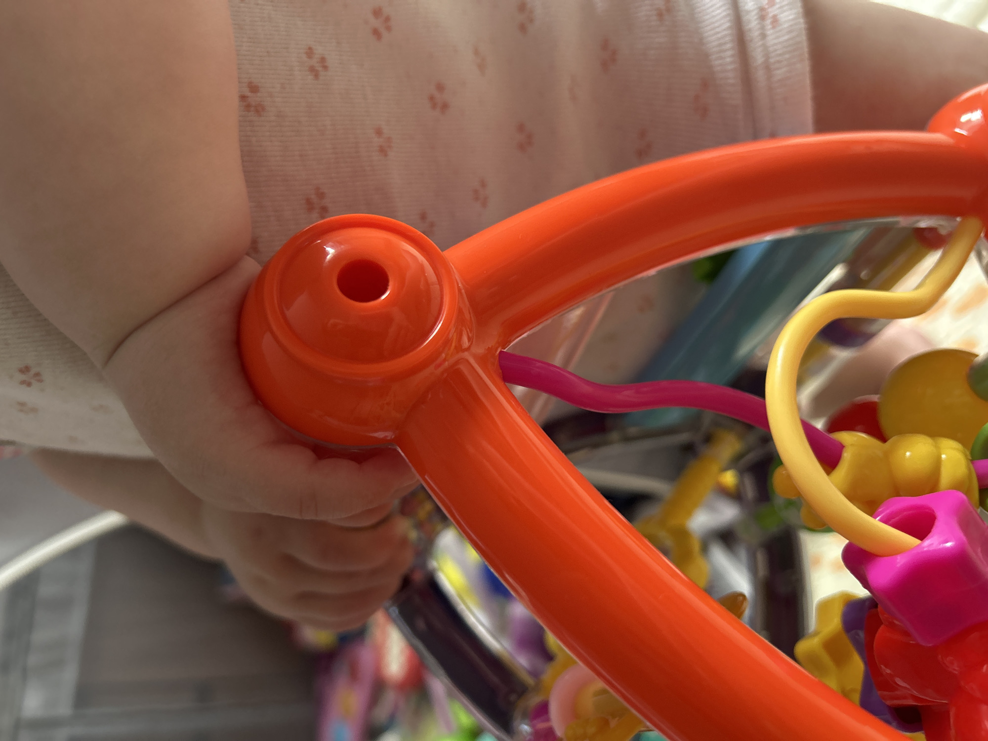 A close-up shot captures a baby's hand grasping an orange plastic toy. The toy features a central ring with a hole in the middle. The baby is wearing a white onesie with a delicate floral pattern. In the background, other colorful baby toys are visible, creating a playful and cluttered environment. The lighting is bright and natural, suggesting an indoor setting, possibly a nursery or play area. The image is focused on the interaction between the baby and the toy, highlighting the tactile experience of childhood play.