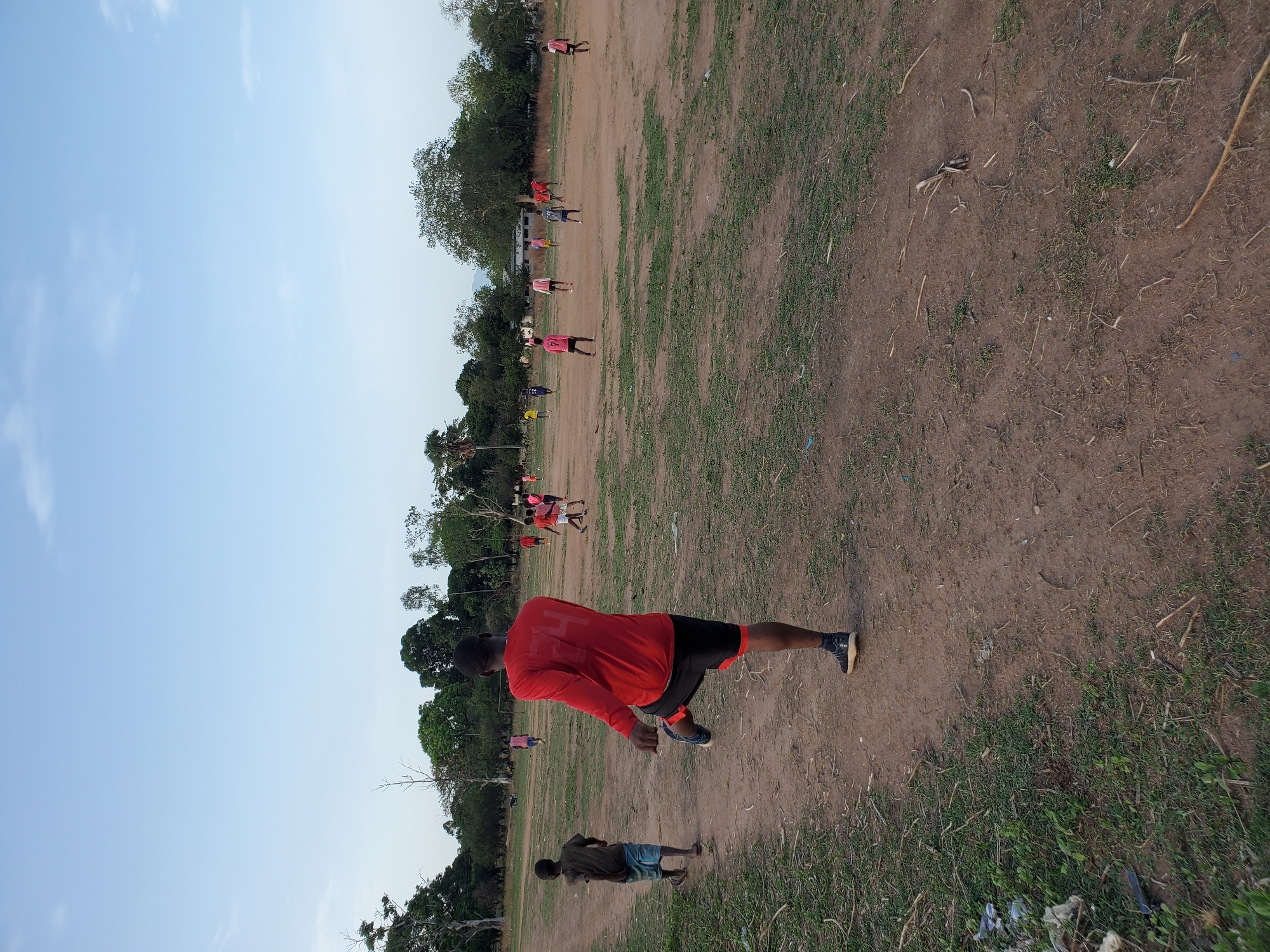 This image depicts an outdoor scene in an open, dry, and dusty field, likely in Mararaba, Nigeria, under a bright blue sky with a few faint clouds. The ground is predominantly light brown earth, interspersed with patches of sparse green grass and some dry vegetation or debris.
In the foreground, a man, viewed from behind, wears a long-sleeved red shirt and black shorts with a red stripe, along with dark sneakers. He is captured in a dynamic pose, leaning forward with his left leg extended backward, suggesting he is in motion, possibly walking briskly or performing an exercise. A faint white marking, resembling a letter or logo, is visible on the back of his red shirt, though it is not clearly legible. To his left and slightly further back, a smaller individual or child, also seen from behind, stands wearing a dark t-shirt and light blue shorts.
Numerous other people are scattered across the mid-ground and background of the field. Many of these individuals are also dressed in red or pink t-shirts or jerseys, indicating they might be part of the same group or team. They appear to be standing, walking, or engaging in various informal activities, possibly preparing for or participating in a sports practice or a community exercise event. Lush green trees line the far edge of the field, creating a natural backdrop. The scene conveys a sense of casual outdoor activity during what appears to be daytime, with clear weather conditions.