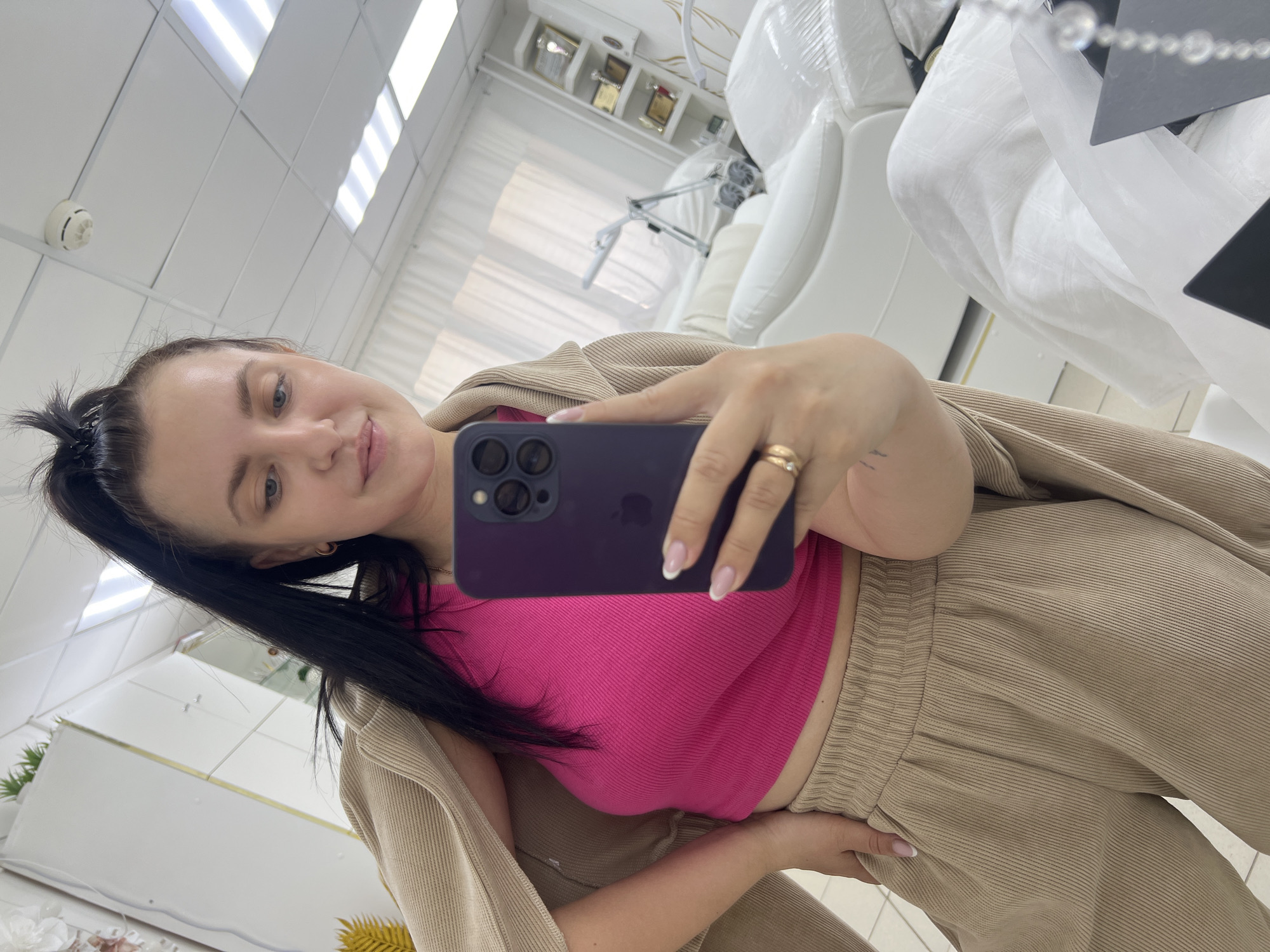 A young woman with dark hair pulled into a high ponytail, wearing a bright pink crop top and beige corduroy pants, takes a selfie in a room that appears to be a beauty salon or spa. She is holding a dark purple smartphone in her right hand. The room features white upholstered furniture, a shelf with awards, and lighting fixtures on the ceiling. The woman is standing in front of a large mirror, as indicated by the reflection of the room in the background.