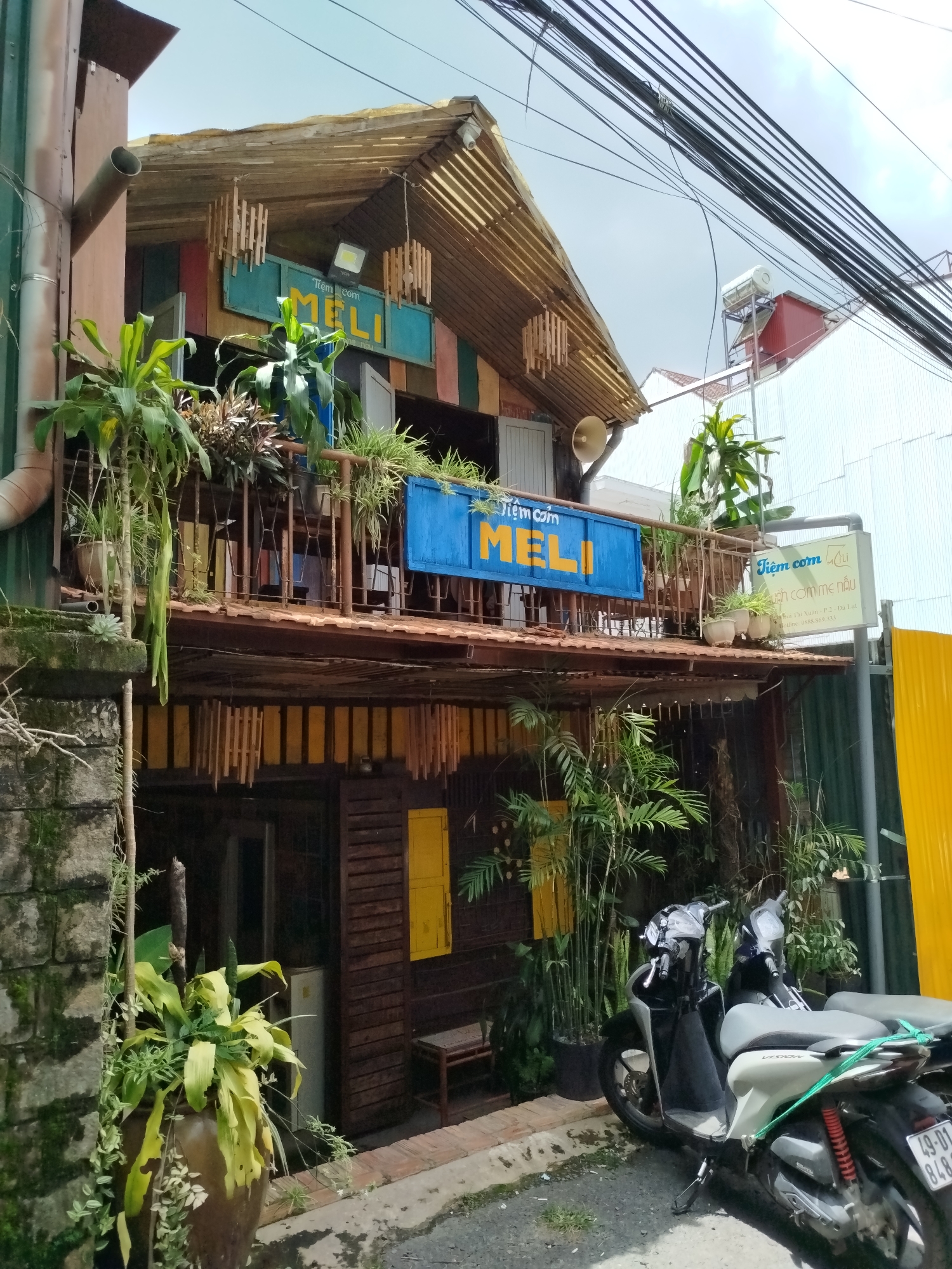 The image captures a street scene in Da Lat, Vietnam, featuring a two-story establishment with a rustic, wooden facade. The building is adorned with colorful wooden panels and decorated with hanging bamboo elements and potted plants. A prominent blue sign with yellow lettering reads "Tiệm cơm Meli," indicating a rice eatery. Another sign to the right displays "Tiệm cơm Meli" and additional Vietnamese text, along with a phone number.
The ground floor has wooden shutters and doors, with a large brown ceramic pot overflowing with green plants positioned to the left. A small, rustic bench is visible outside the ground floor entrance.
The upper floor features a balcony area, also decorated with plants, with a railing constructed from metal bars. Two scooters are parked in front of the building, one light-colored and the other dark, with a green rope tied around the handlebars of the light-colored scooter. The environment appears to be an urban setting with power lines crisscrossing overhead. The sky is overcast.