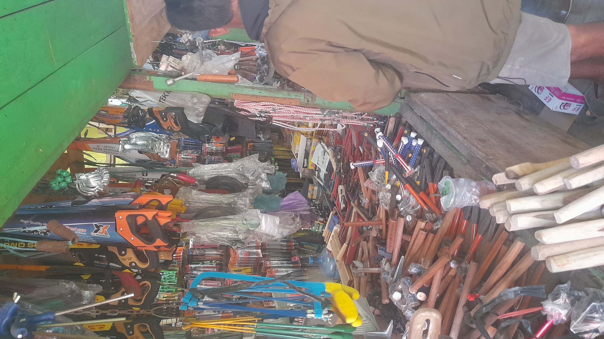 The image shows a street-side hardware stall in Pekalongan, Indonesia. The stall is filled with a variety of tools, including saws, hammers, and other hand tools. The tools are displayed on shelves and in bins, with some items hanging from the ceiling. A man in a brown jacket and light-colored shorts is standing in the foreground, facing away from the camera. The stall appears to be open to the street, with a green awning providing shade. The overall impression is of a busy and well-stocked market stall.