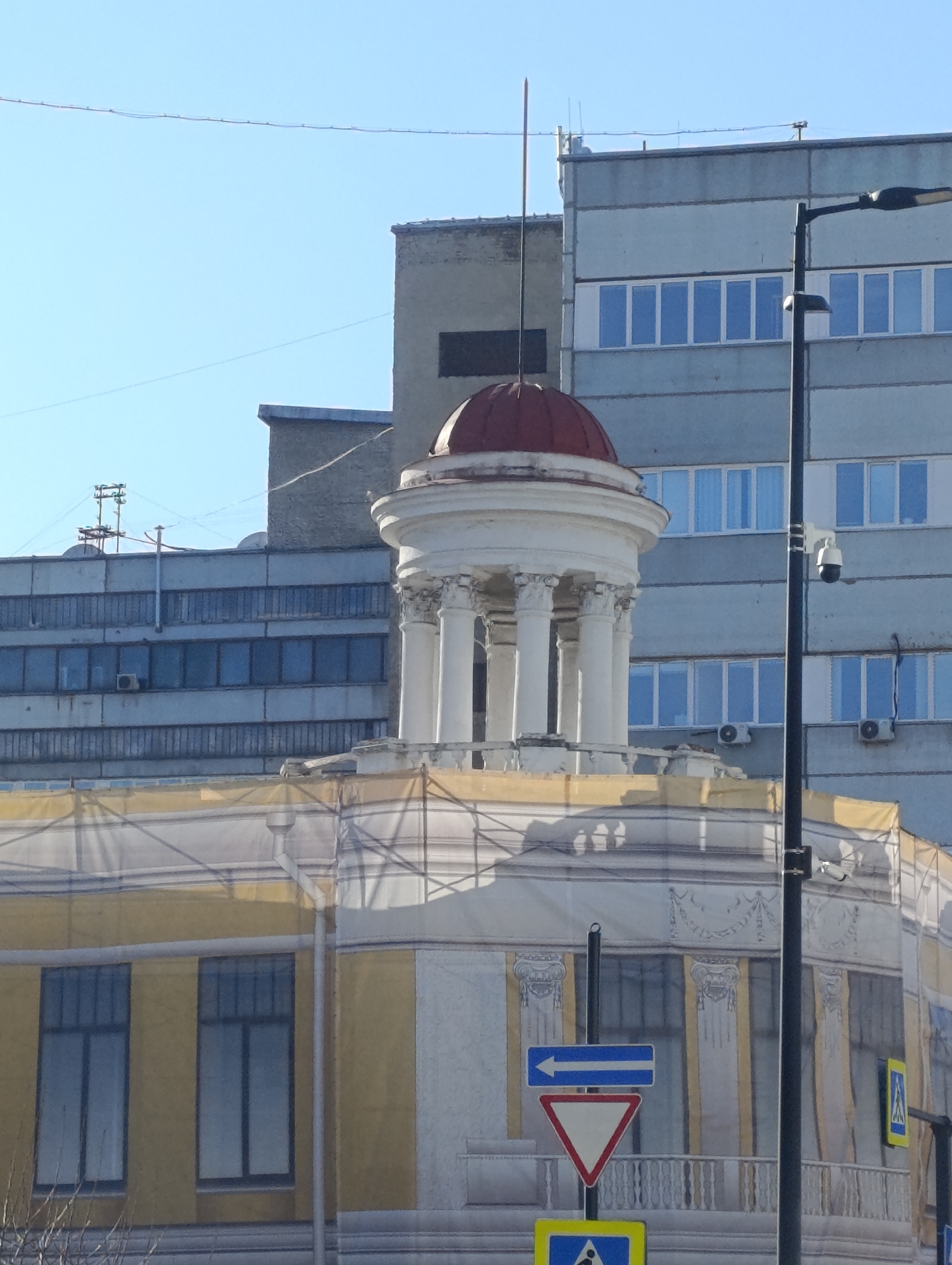 The image shows a building in Krasnoyarsk, Russia, undergoing renovation. A yellow and white building is partially covered with a tarp or scrim featuring an image of the building's original facade. Above the main building is a red-domed rotunda supported by white columns. Behind the building, are two grey buildings with rows of windows. A black street lamp is situated to the right of the building. Street signs, including a left arrow and yield sign, are visible at the base of the building. Bare tree branches are visible in the foreground. The sky is clear and blue.