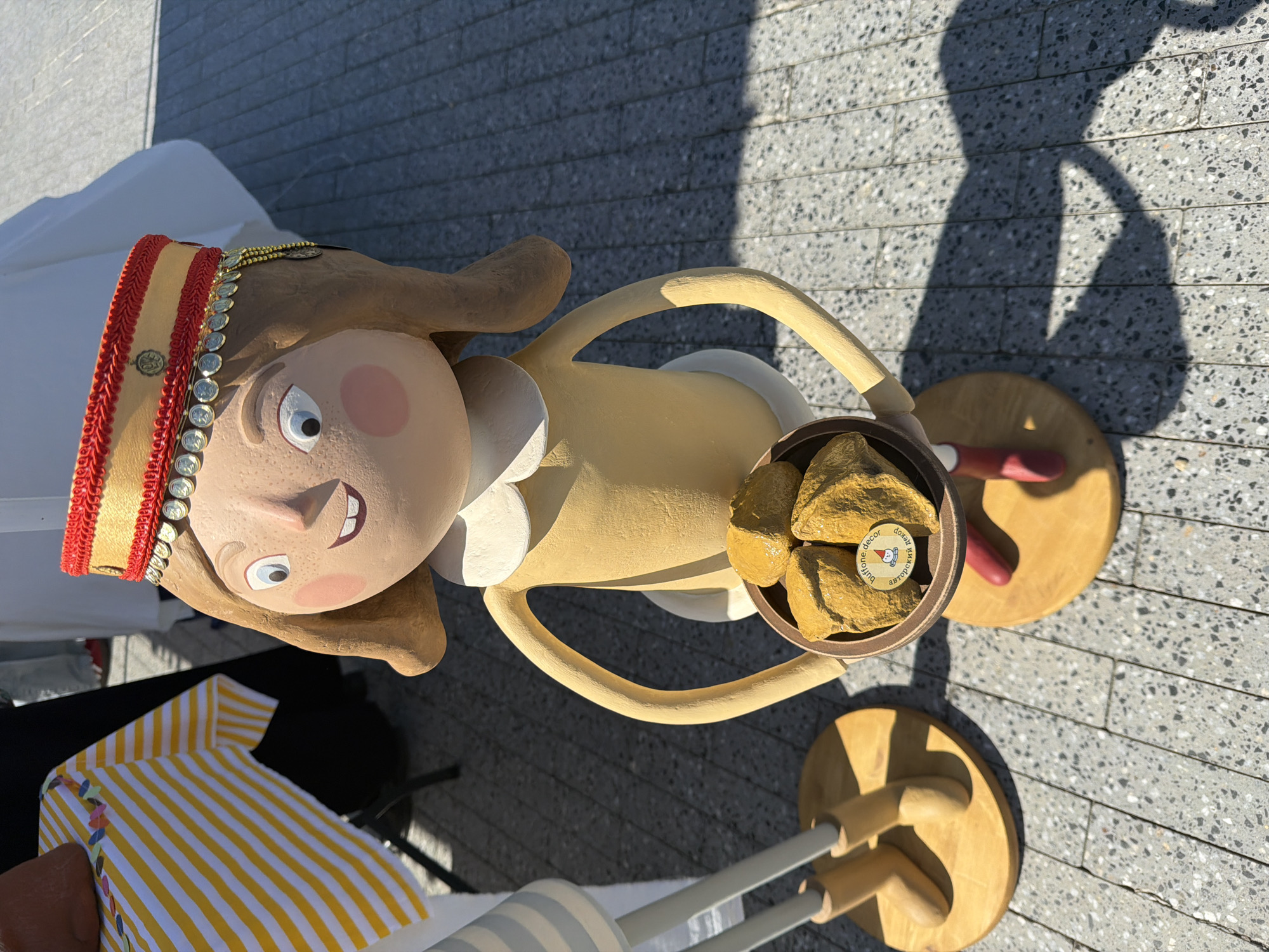 A full shot shows a life-sized doll with a yellow dress, red shoes, and a red and gold hat with coins. The doll is standing on a wooden base with a bowl of golden rocks in its hands. The doll is in front of a table with a yellow and white striped cloth. The background shows a paved area with shadows. The scene is outdoors, likely in Kazan, Russia.