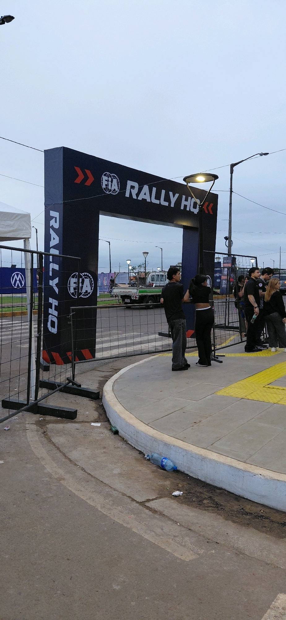 The image depicts an outdoor scene in Encarnacion, Paraguay, under an overcast sky. In the foreground, a black archway bearing the "FIA Rally HQ" logo stands prominently, marking an entrance or checkpoint. The archway has red chevron graphics on its sides and a light fixture above. To the left of the archway, a temporary white tent structure is partially visible, along with a black metal fence.
On the paved walkway in front of the archway, a group of people are gathered. Two individuals, dressed in dark clothing, appear to be interacting. One, a person with dark hair wearing a black top and pants, is facing away from the camera and seems to be speaking to another person in a black t-shirt and jeans, who is also facing away. Further to the right, several other individuals, also dressed in dark attire, are standing near the archway.
Behind the archway and the people, a light-colored truck with "FIA MOTORS" branding on its side is visible, parked on a road. More temporary fencing and banners with logos are present in the background, suggesting a public event or gathering. The ground in the immediate foreground and along the curb is dirty, with litter including a discarded plastic water bottle. The overall environment appears to be a public space adapted for an event.