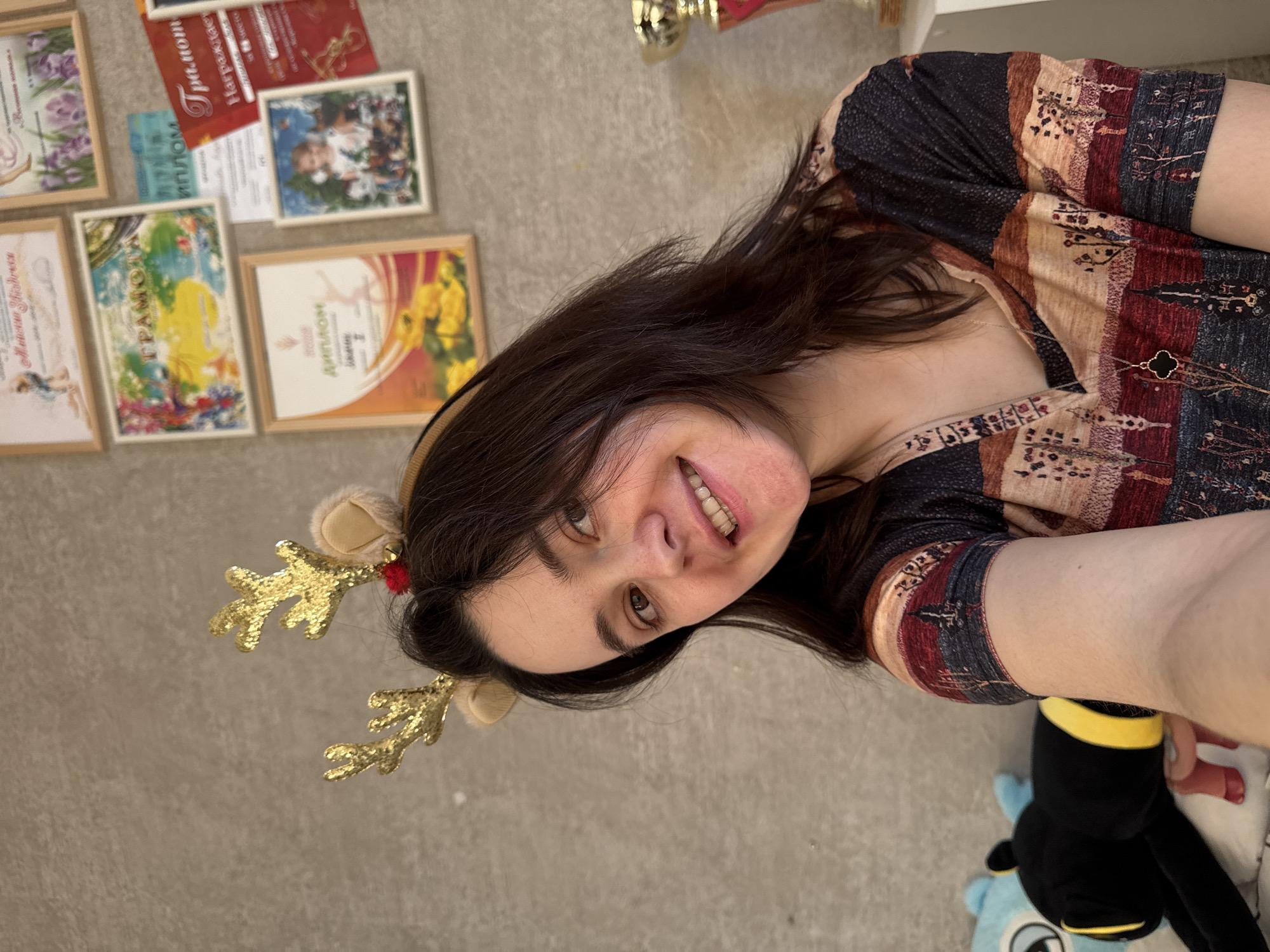 A woman with dark hair is lying on a light beige floor wearing a patterned shirt and reindeer antlers with gold glittery antlers. She is smiling and looking at the camera. On the floor next to her are several framed certificates and awards. A stuffed animal toy is to the bottom right of her. The photo was likely taken indoors but no location of Izhevsk, Russia can be definitively stated based on the available information.