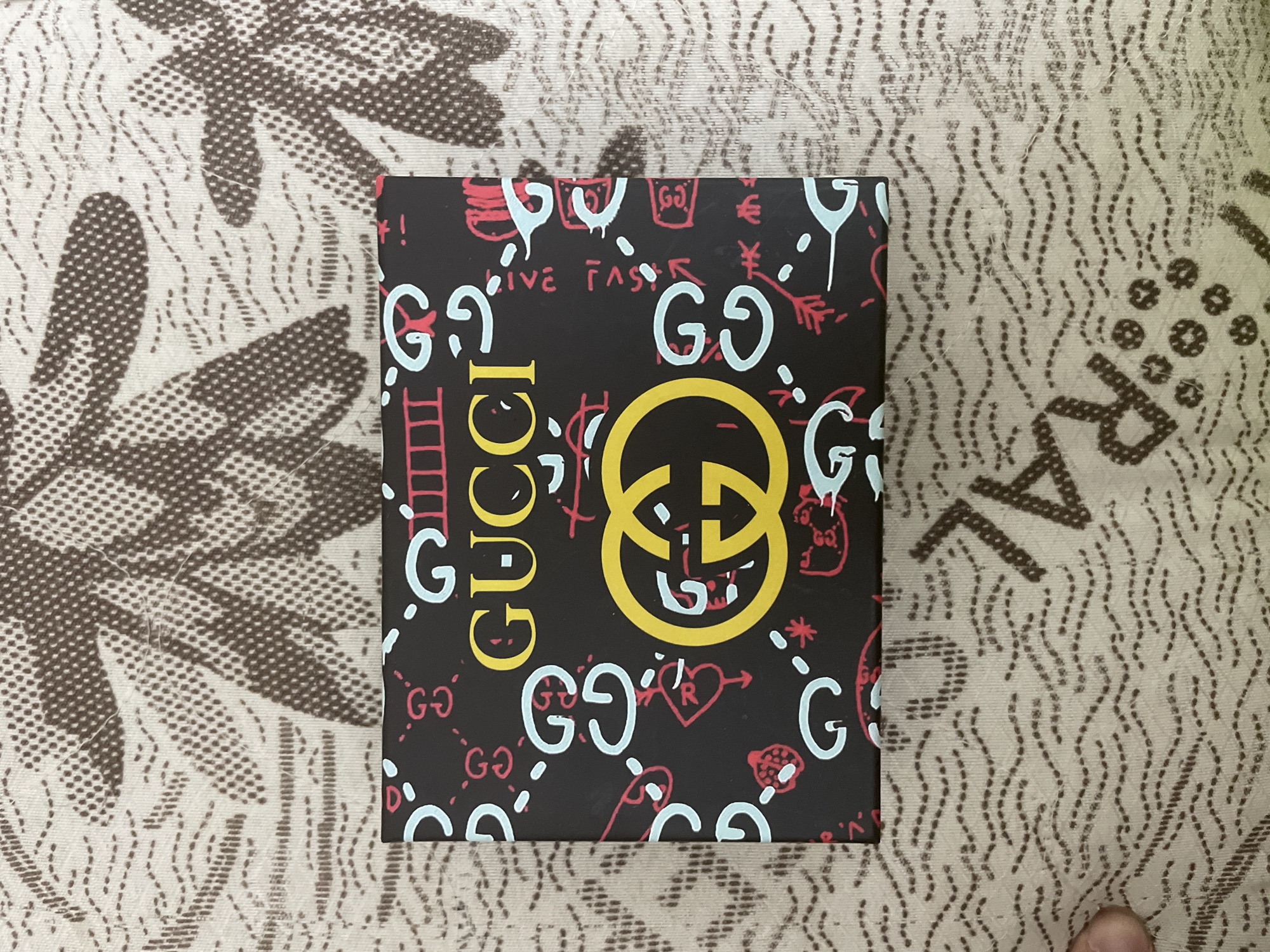 The image presents a rectangular, dark-colored Gucci product box positioned centrally on a light-colored, patterned fabric surface. This scene is captured indoors, likely in Ihtiman, Bulgaria.
The box features a vibrant, graffiti-inspired design. Prominent yellow text on the left side vertically spells "GUCCI," while a large, yellow interlocking "GG" logo, the brand's iconic symbol, dominates the center. The box's dark background is filled with numerous doodle-like graphics and text in contrasting light blue/cyan and red. Light blue elements include multiple stylized instances of "GO" and individual "G" letters. Red elements comprise various small drawings such as hearts, arrows, ladders, crowns, what appear to be currency symbols (€, ¥), and other abstract shapes. Partial red text spelling "LIVE FAS" is also visible, strongly suggesting the phrase "LIVE FAST."
The fabric beneath the box is a light beige or off-white color and displays a repeating dark brown pattern. This pattern is composed of dense clusters of small dots and flowing dashed lines, creating an organic, possibly floral or wave-like texture. In the bottom right corner of the original image, a human thumb is partially visible, indicating that the box is being held or has just been placed. The lighting is soft and even, typical of an indoor environment. The image emphasizes the box's distinctive and modern aesthetic, showcasing the luxury brand's playful packaging design.