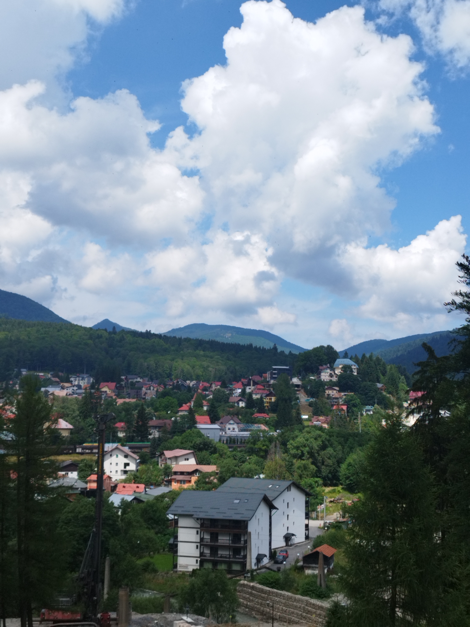 The image depicts a panoramic view of the town of Bușteni, Romania, nestled at the foot of the Bucegi Mountains. The scene is dominated by lush green forests covering the mountainsides, with scattered houses and buildings forming the town below. The sky is a vibrant blue, punctuated by fluffy white clouds. In the foreground, there's a modern multi-story building with a dark roof and white walls, featuring balconies. To the left of this building, a large drilling rig is visible, suggesting ongoing construction or infrastructure work. The town itself is characterized by a variety of residential buildings, many with red or orange roofs, interspersed with trees. The perspective suggests the photograph was taken from a slightly elevated position, looking down on the town and towards the mountains. There are no people clearly visible in the image. The overall impression is one of a picturesque mountain resort town during daytime.