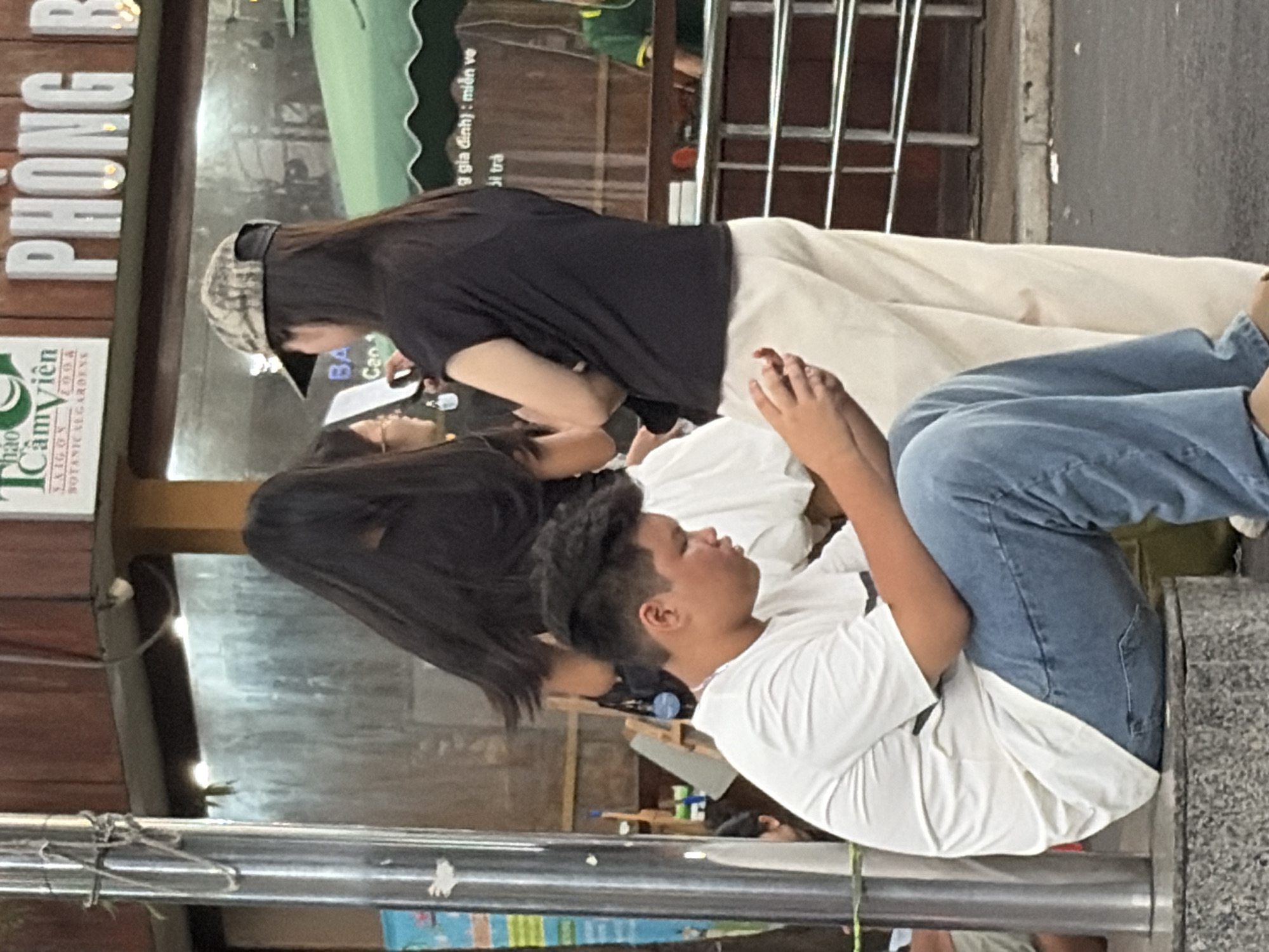The scene takes place in the city of Thủ Đức, Vietnam. An outdoor urban environment is depicted, featuring five visible individuals.
In the immediate foreground, a male individual is seated on a grey, polished stone ledge. He possesses dark, short hair and wears a white short-sleeved t-shirt and blue denim jeans. Both hands are engaged with a black smartphone, to which his gaze is directed downwards.
In the midground, to the right of the seated male, two females are standing. The female closer to the center has long dark hair and wears a black t-shirt paired with light-colored, possibly beige or white, loose-fitting trousers. A camouflage-patterned baseball cap is worn backward on her head. She holds a dark-colored smartphone in her left hand, with her attention seemingly directed towards it or an object to her left. Adjacent to her, a second female stands with her back partially to the viewer, also possessing long dark hair and wearing a black t-shirt. She appears to be interacting with the first standing female. Partially obscured behind the seated male's left side, a leg and arm of another individual wearing a white garment are visible.
In the background, a commercial setting is visible. A prominent white and green sign with "THẢO CẦM VIÊN SAIGON BOTANICAL GARDENS" in English and Vietnamese is mounted on a building facade. Below this, large white raised letters spell out "PHÒNG B...". A green awning extends over a counter on the right side of the image, behind which a person wearing a green shirt is partially visible, suggesting an establishment. The background also features large glass panels reflecting ambient light and structures, along with a metal railing system.
A tall, polished metal pole with various wires or ropes wrapped around it stands on the far left. The ground is asphalt, with some minor debris visible. The overall lighting suggests daylight, possibly late afternoon, with artificial lights illuminating the background commercial areas.
