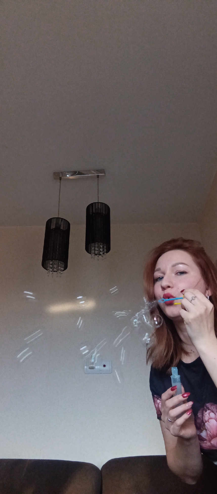 A woman with red hair is blowing bubbles indoors. She is holding a blue and yellow bubble wand to her lips, and there are several bubbles floating in the air in front of her. She is also holding a small blue bottle in her other hand. She has red nail polish and rings on her fingers. Behind her is a white wall with a light switch and a hanging light fixture with two black shades. A dark couch is visible at the bottom of the frame. The location is Unknown, Unknown.