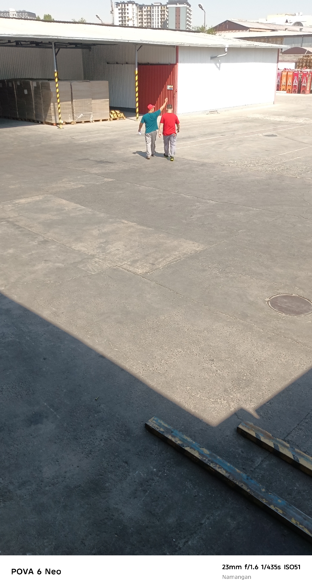 The image depicts an outdoor scene in Namangan City, Uzbekistan, likely an industrial or storage area. In the mid-ground, two individuals are walking away from the camera on a paved surface. One person is wearing a blue shirt and gray pants, and the other is wearing a red shirt and gray pants. The person in the blue shirt has their right arm raised. Behind them is a large, white building with a red door and a covered awning. Stacks of what appear to be cardboard boxes or similar materials are visible under the awning to the left. In the foreground, a large shadow is cast diagonally across the pavement from the left, obscuring some of the surface. Two metallic objects with yellow and blue markings lie on the ground in the lower right portion of the image. The background shows buildings, including some tall structures that suggest a city environment. The lighting indicates it is daytime, with clear sky visible.