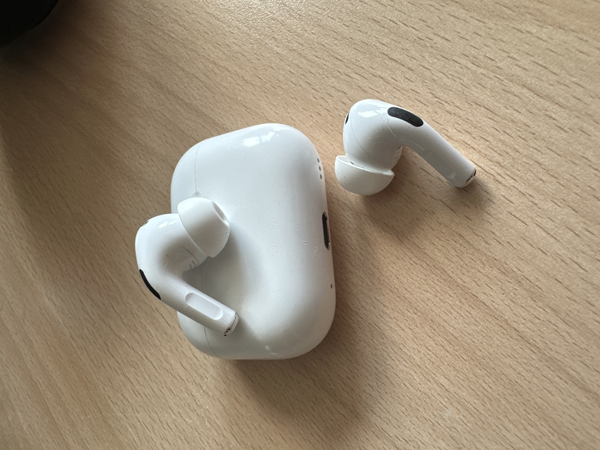 A white charging case for wireless earbuds rests on a wooden surface. One wireless earbud is partially inserted into the case, while another is lying separately to the right of the case. The wooden surface has a visible grain pattern.